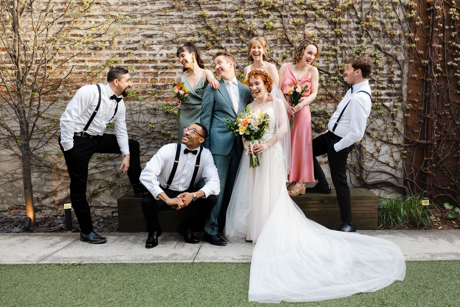 Bride and groom pose with wedding party against brick wall covered in climbing vines.
