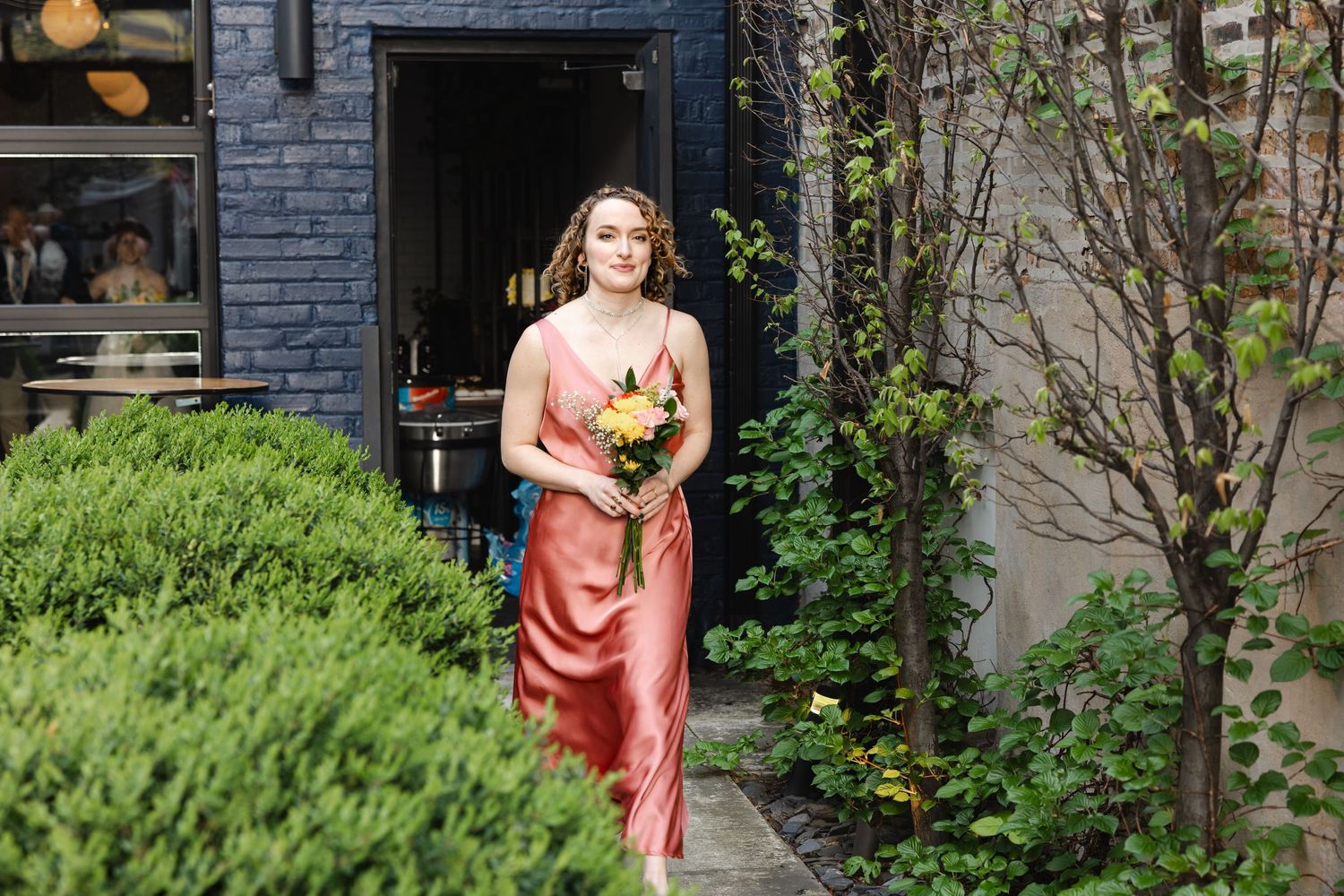 Woman in coral satin dress holding colorful bouquet walks through lush garden pathway near brick building.