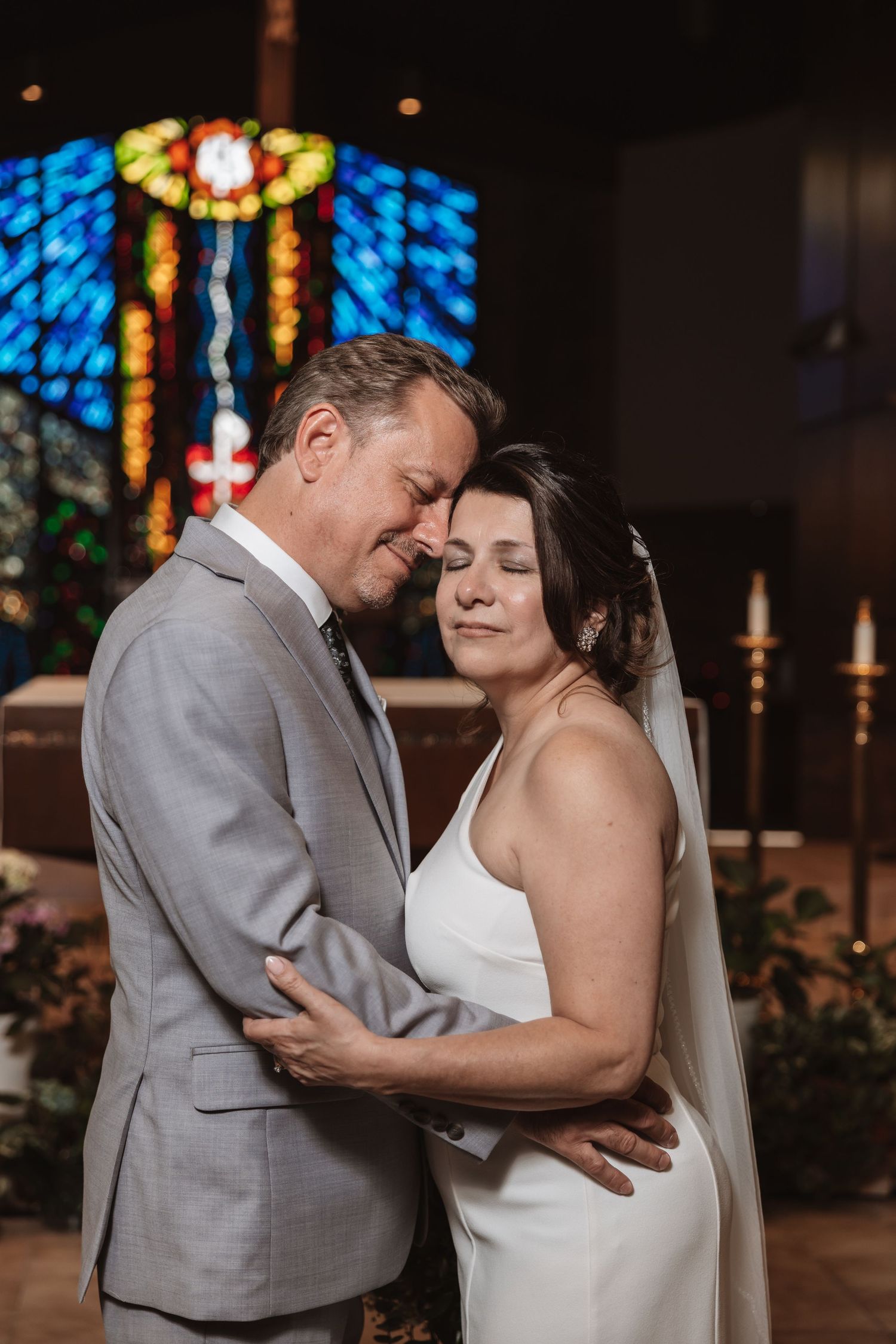 Bride and groom share tender forehead touch embrace in church with vibrant stained glass window backdrop.