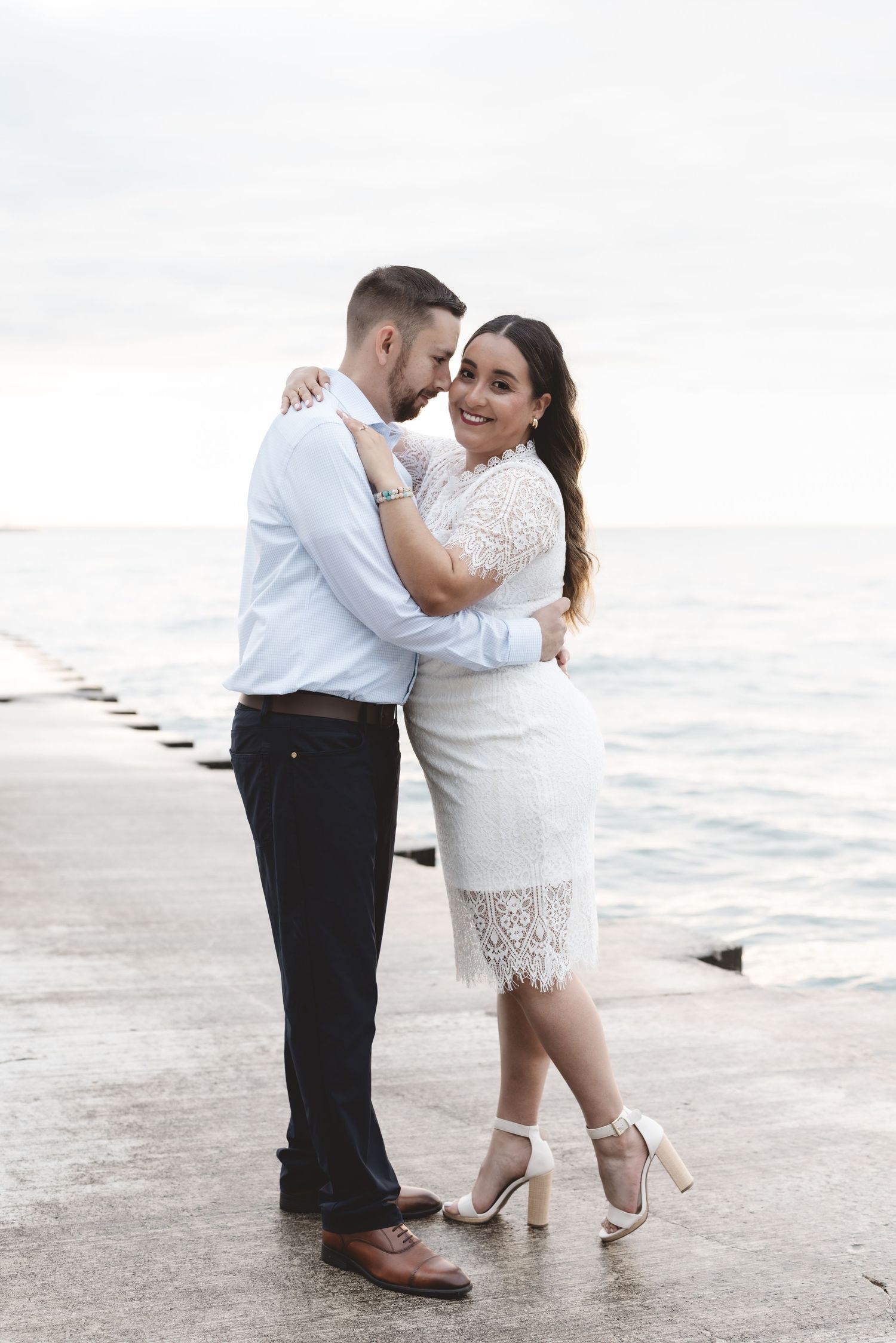 Couple embracing on lakeside pier, woman in white lace dress and heels smiling at camera, man in white shirt and dark pants.