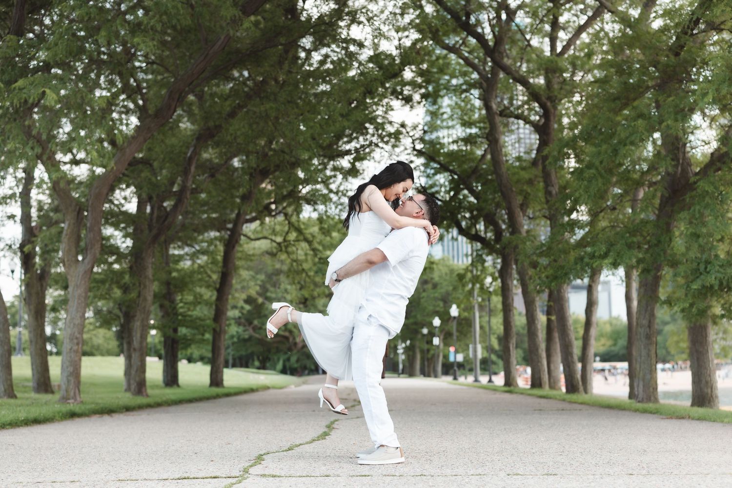 Couple in white outfits embracing joyfully on a tree-lined park path during an outdoor engagement session.