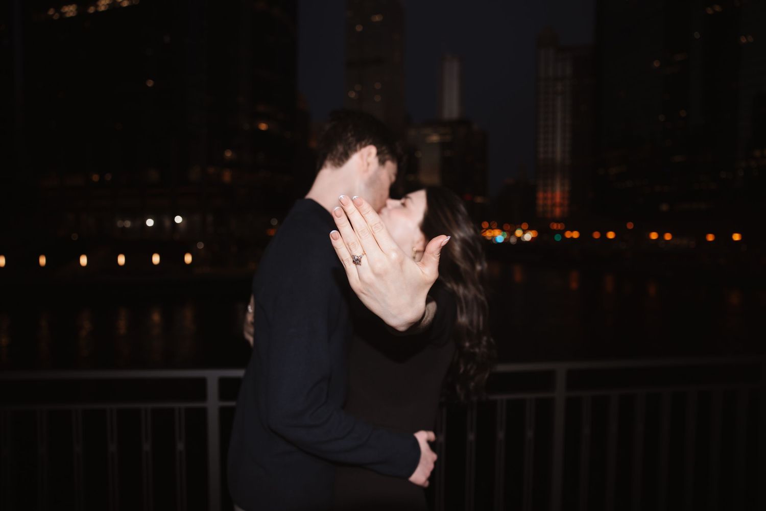 Couple shares a kiss at night by the Chicago River, woman shows off engagement ring with city lights glowing behind them.
