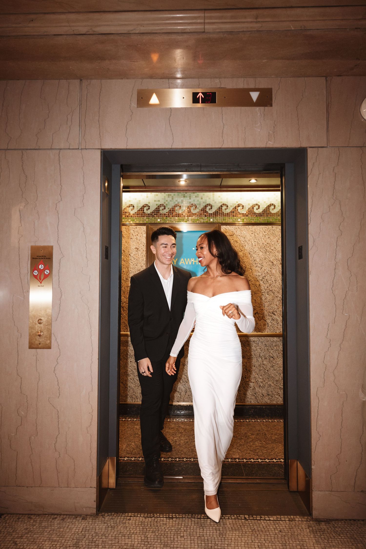 Smiling bride in off-shoulder white gown and groom stepping out of an elegant elevator.