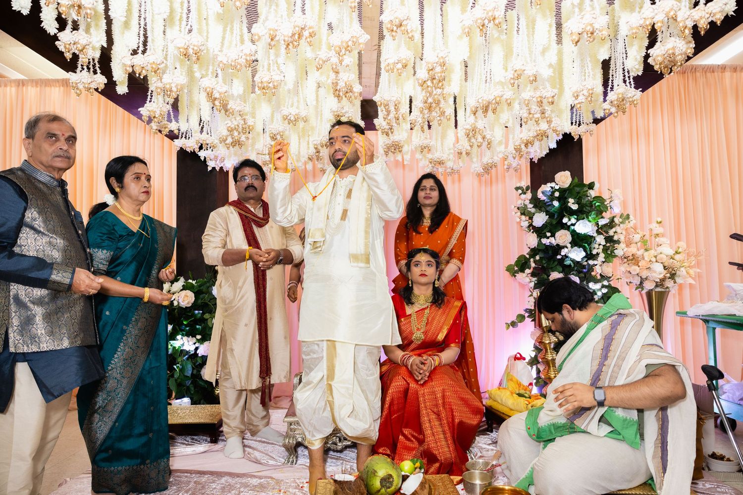Indian wedding ceremony under floral chandelier with bride in red saree seated and guests in traditional attire.