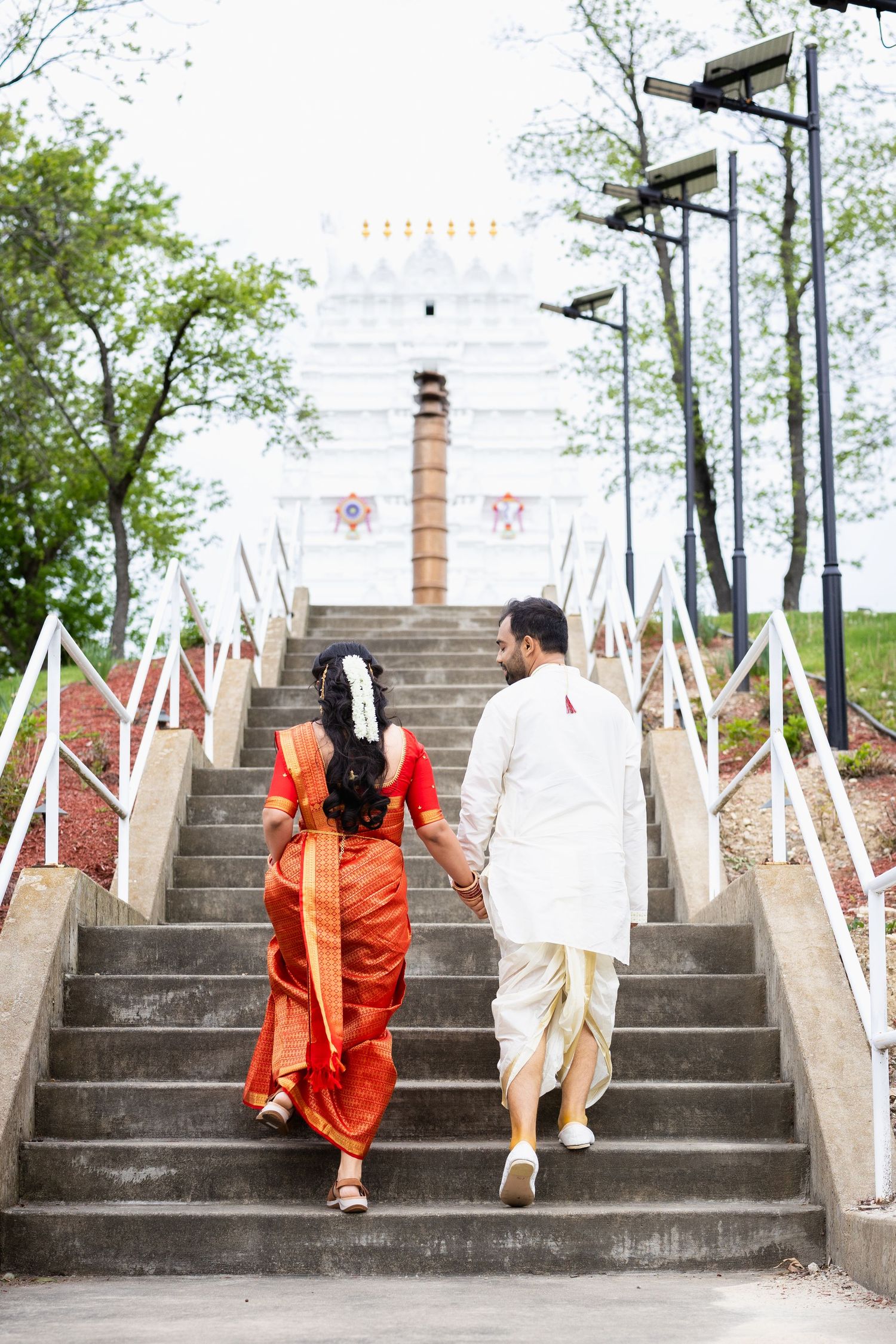 Hindu wedding couple holding hands ascending temple stairs, bride in orange saree and groom in white dhoti.