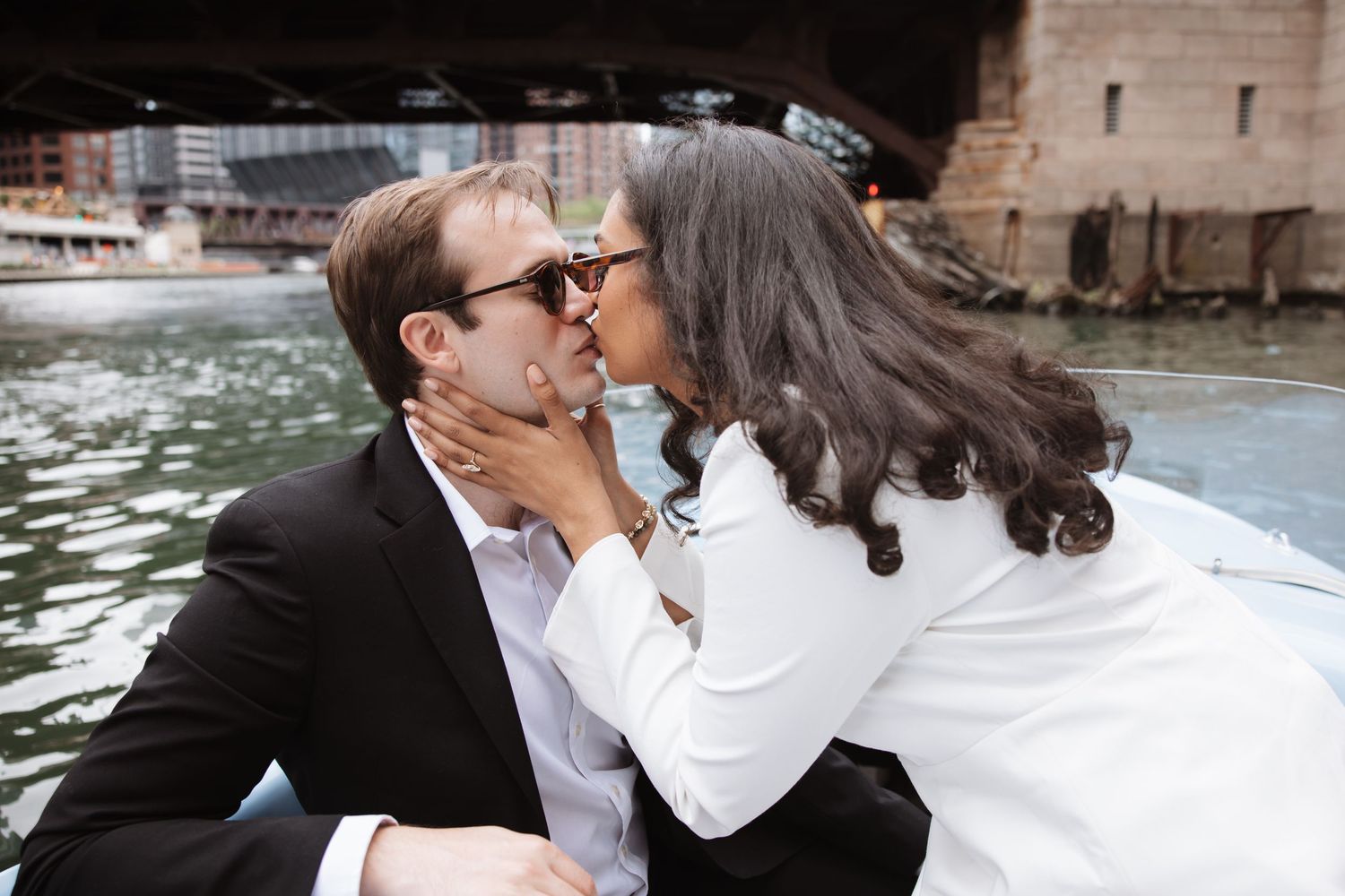 Couple sharing a romantic kiss on a boat under a bridge on the Chicago River, woman in white jacket and man in black suit.