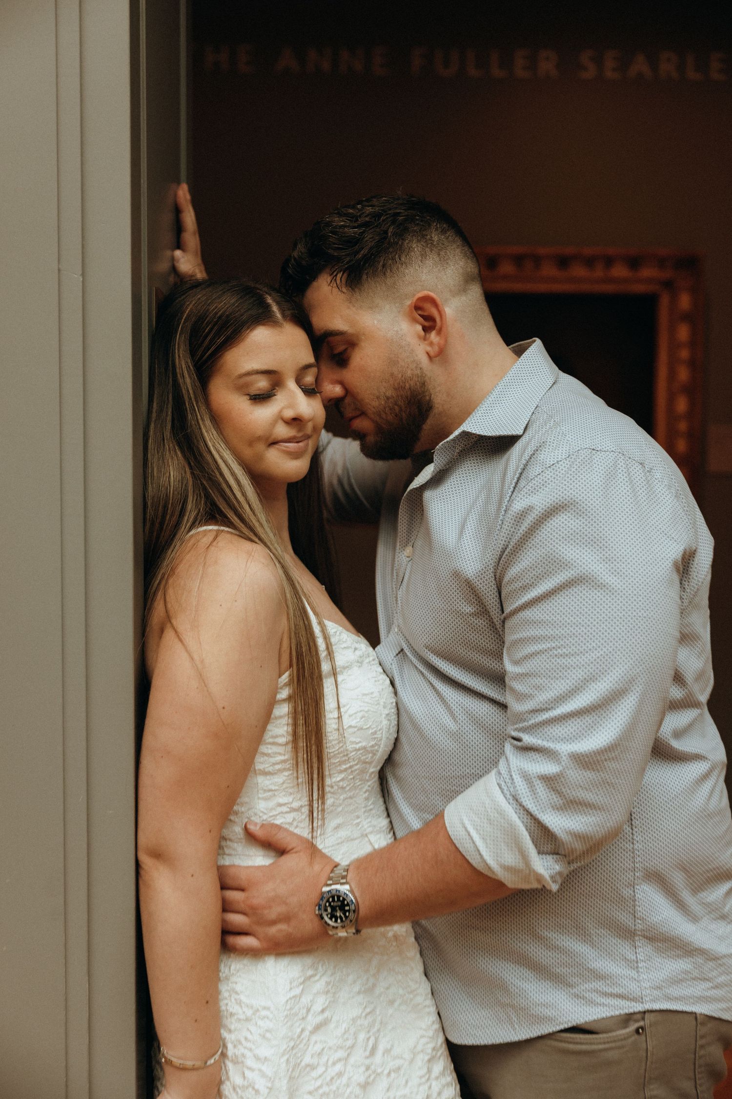 Romantic couple sharing an intimate moment in a museum doorway, woman in white dress, man in blue shirt wearing a watch.