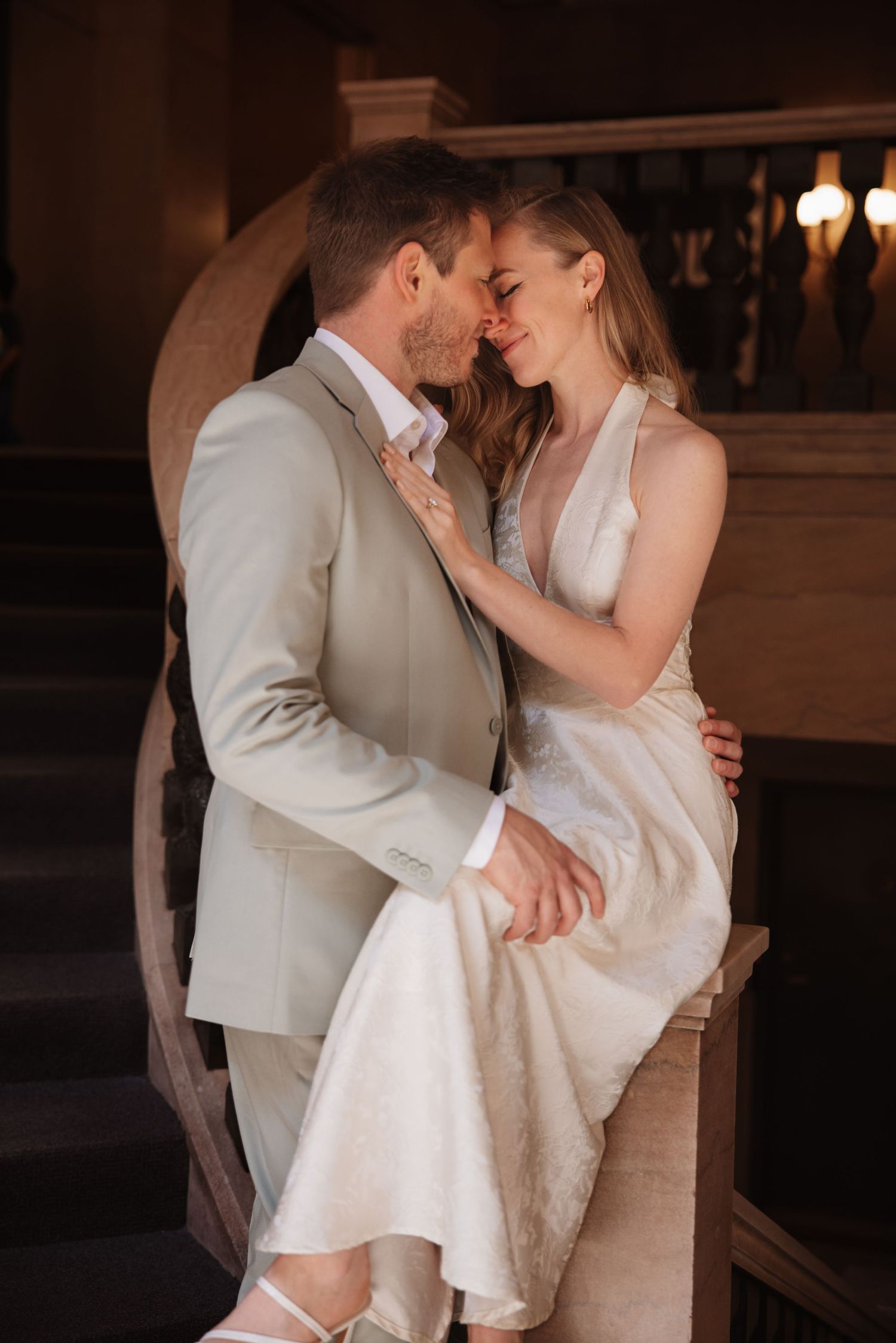 Bride and groom sharing an intimate moment on elegant staircase, she in white gown, he in beige suit.