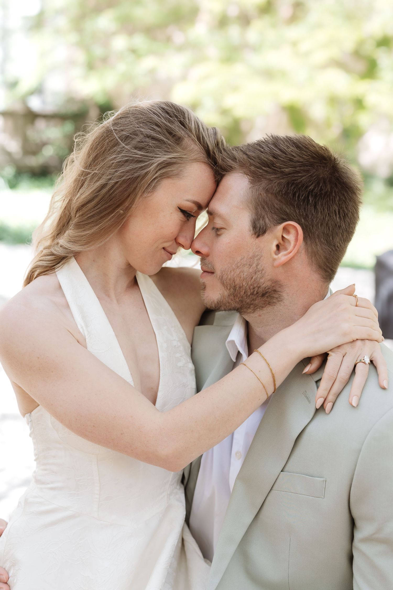 Bride and groom touch foreheads tenderly outdoors, with the bride's diamond ring visible on his shoulder.