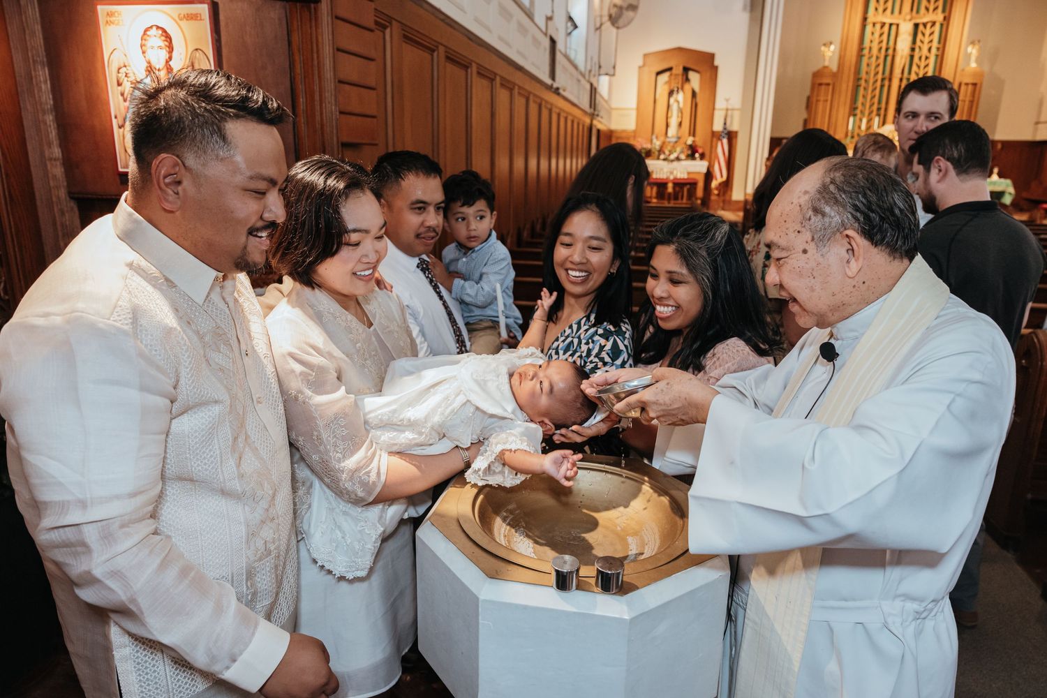 Filipino family and priest gather joyfully around a golden baptismal font during a Catholic christening.