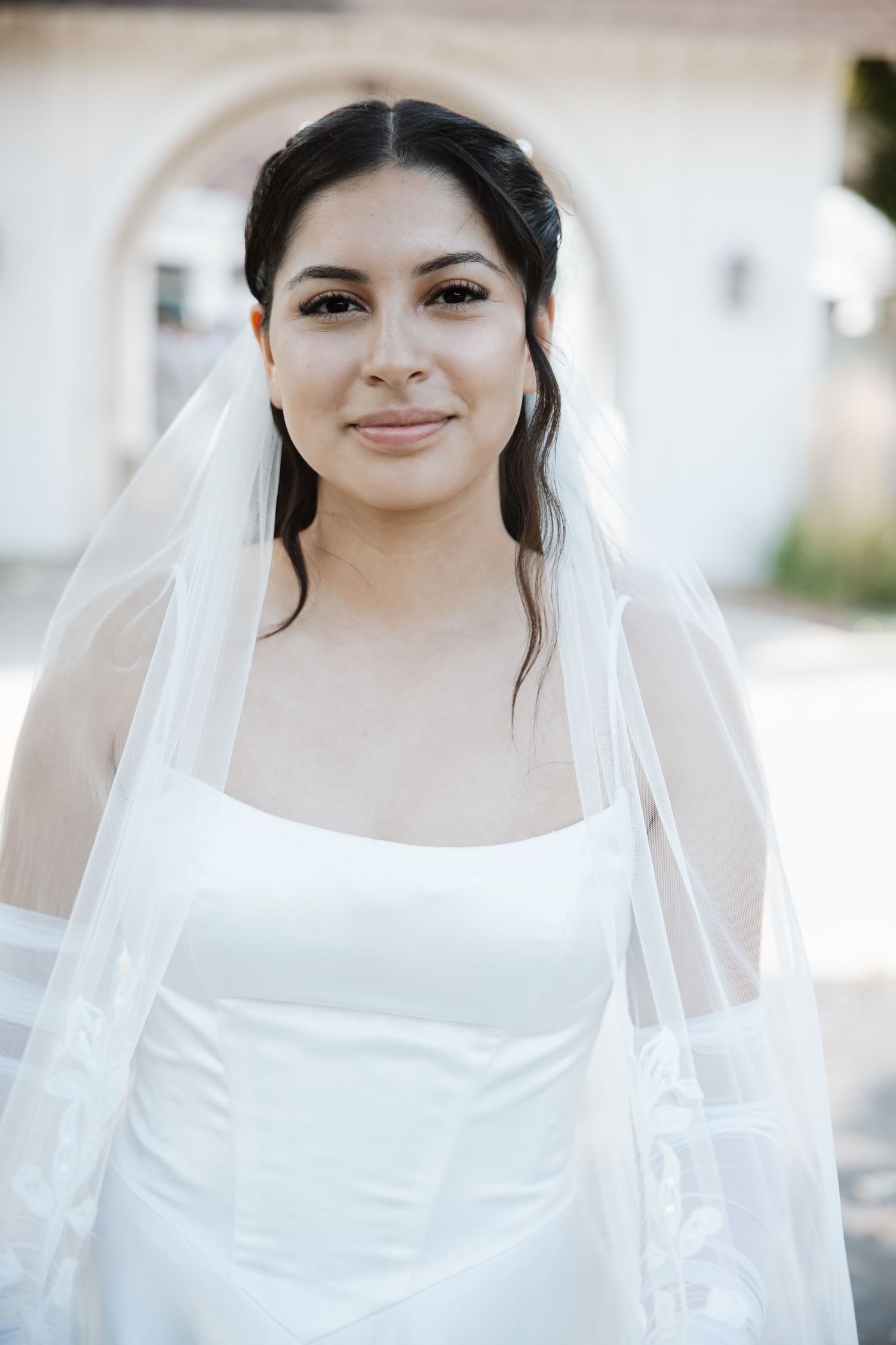 Radiant bride in white gown and sheer veil smiling softly outside white arched venue.