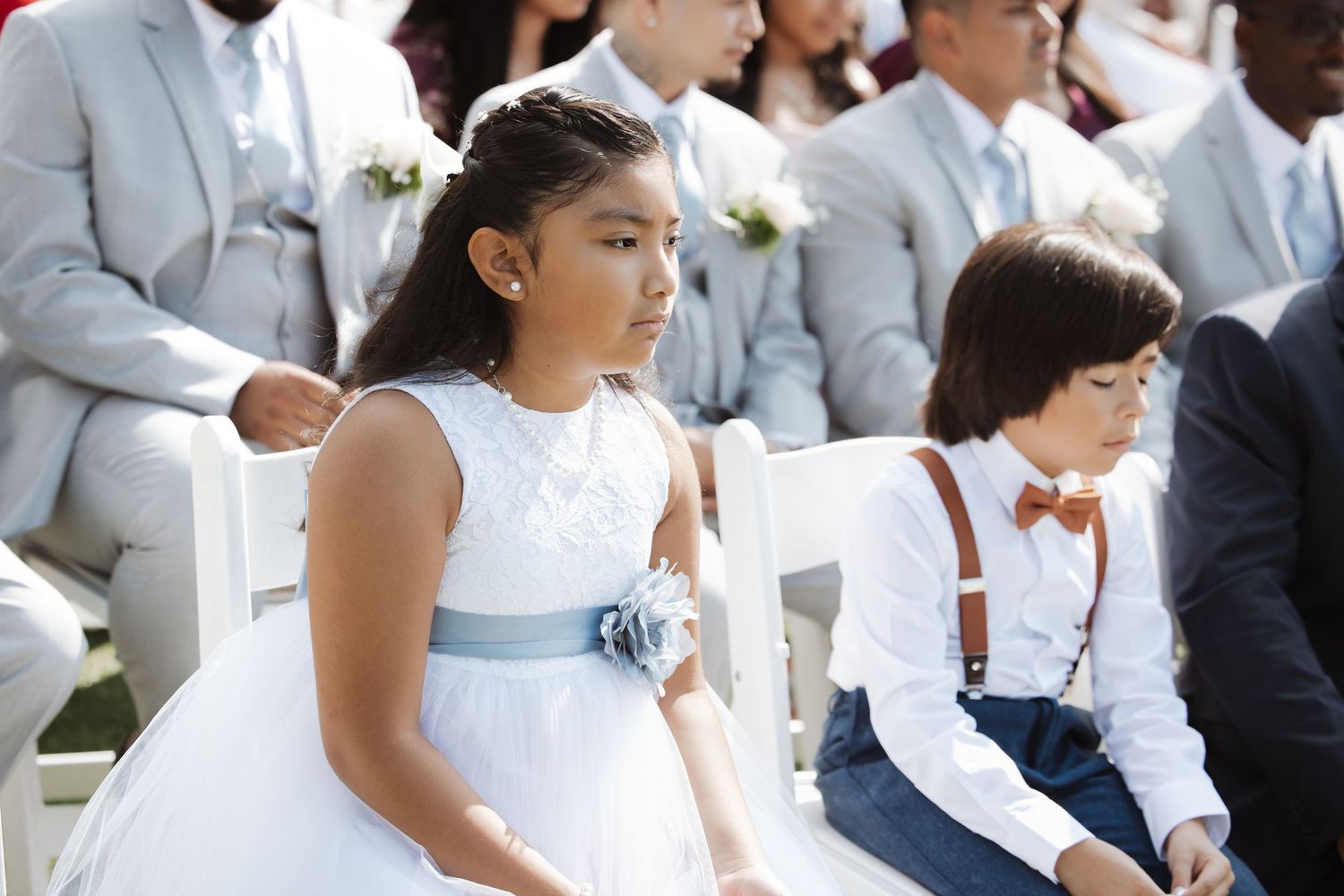 Young flower girl in white dress with blue sash and ring bearer in suspenders at wedding ceremony.