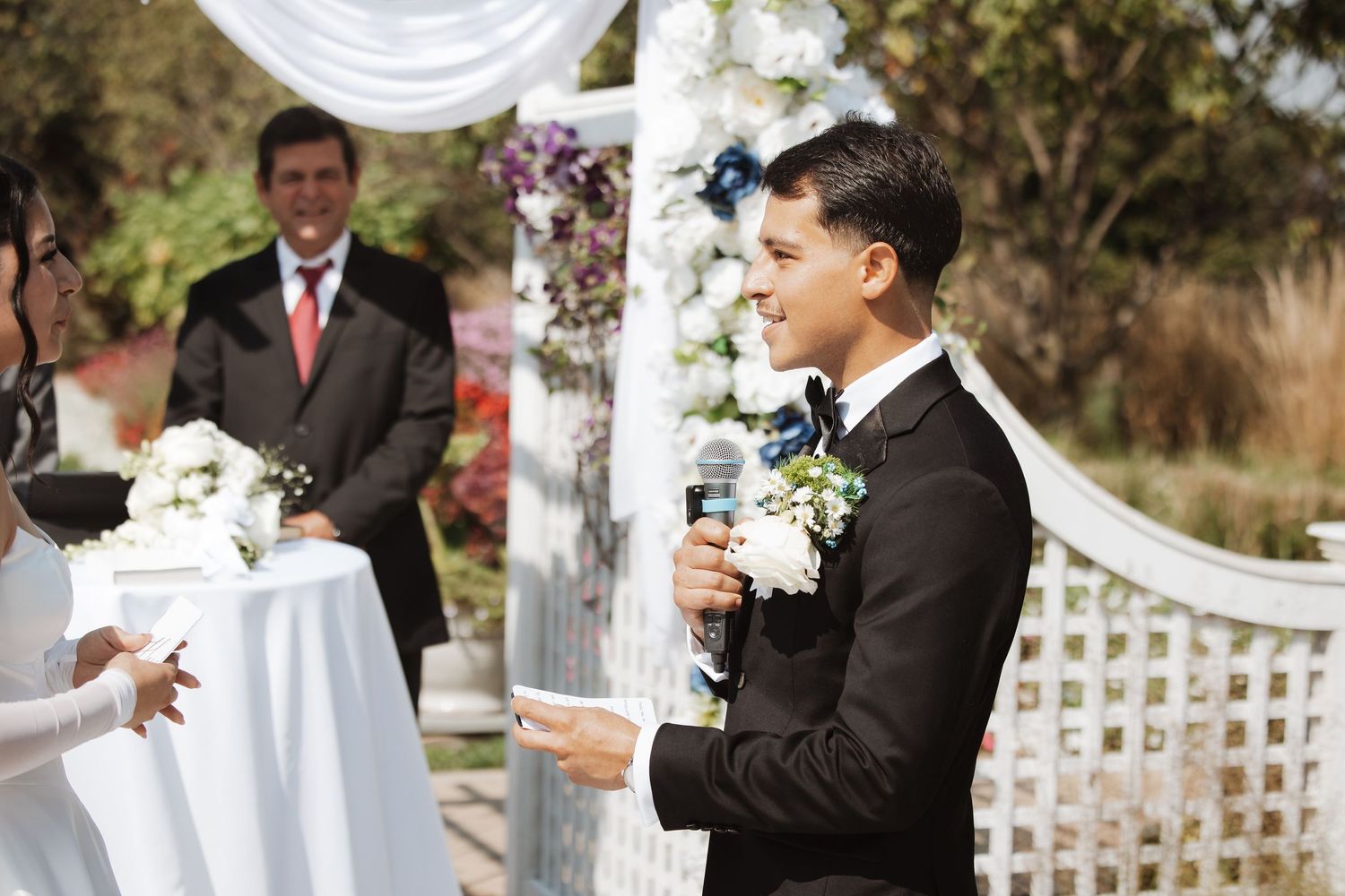 Groom speaking vows into microphone at outdoor wedding altar decorated with white flowers.