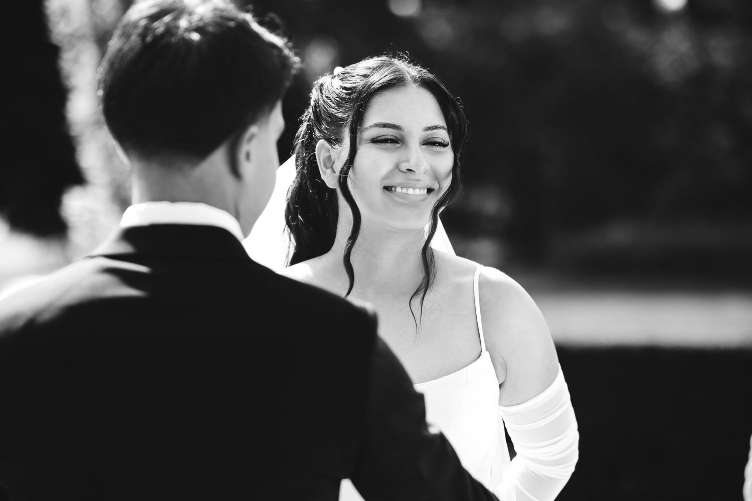 Bride smiling radiantly at groom during outdoor wedding ceremony in black and white photo.