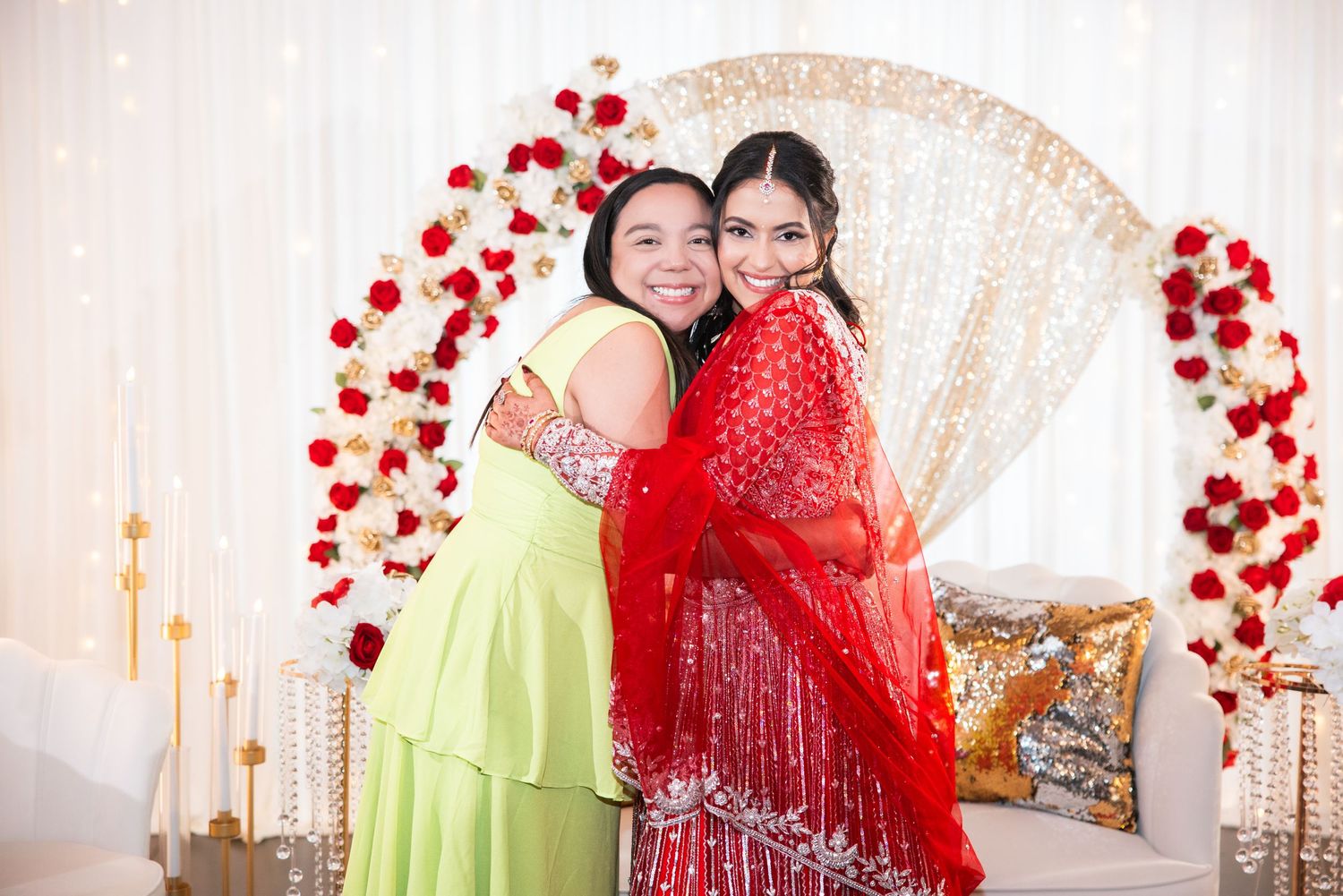 Two women in colorful traditional South Asian attire embracing and smiling at an elegant floral event backdrop.