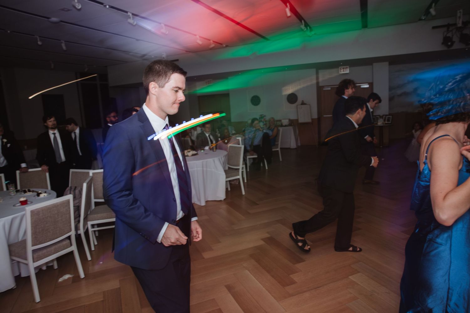 Guest in navy suit dancing at wedding reception with colorful party lights.