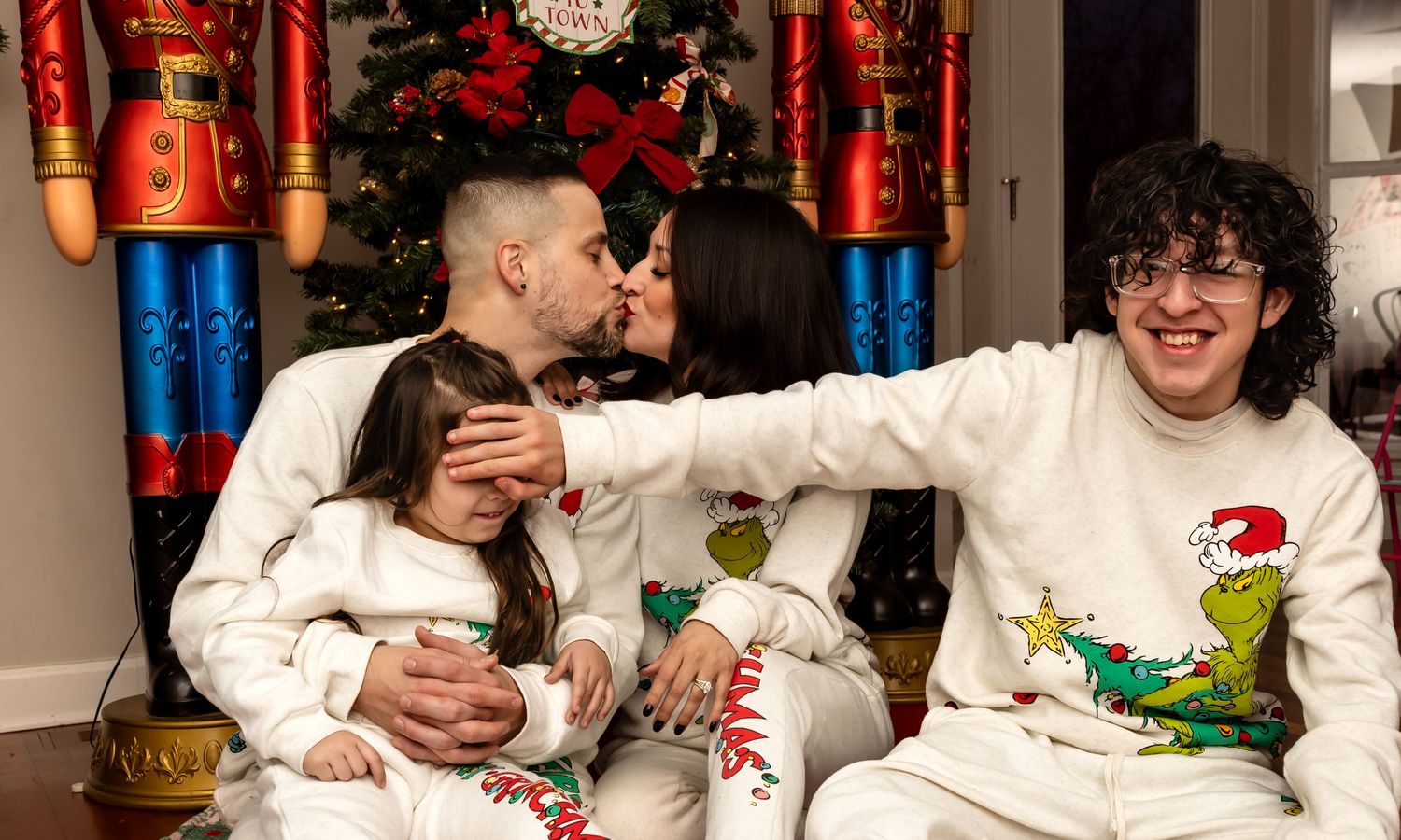 Family wearing matching Grinch holiday pajamas with parents sharing kiss by Christmas tree.