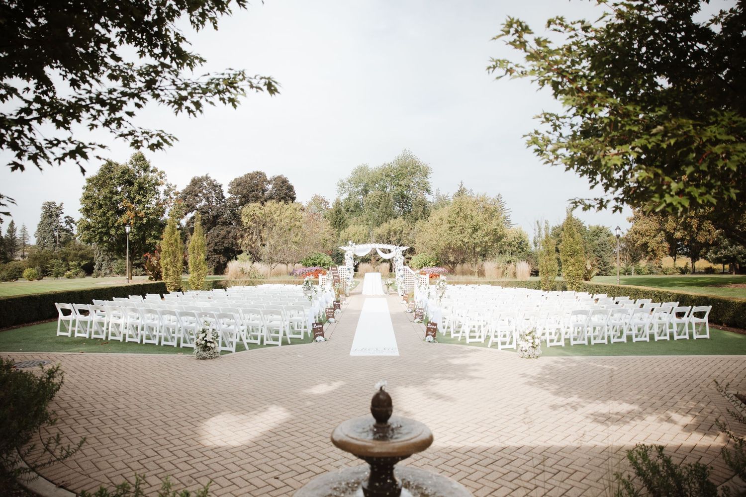 Outdoor wedding ceremony setup with white chairs, floral arch, and brick pathway in a garden.