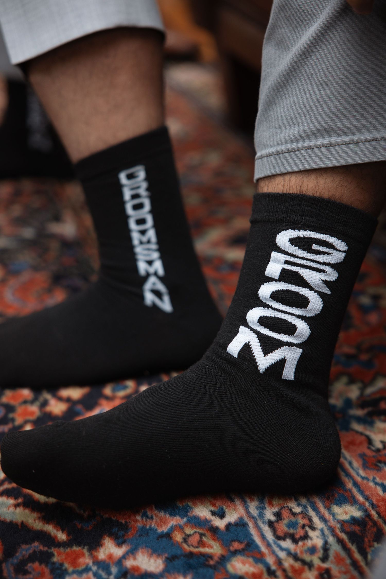 Groom wearing matching black socks with white GROOM text on colorful oriental rug before wedding ceremony.