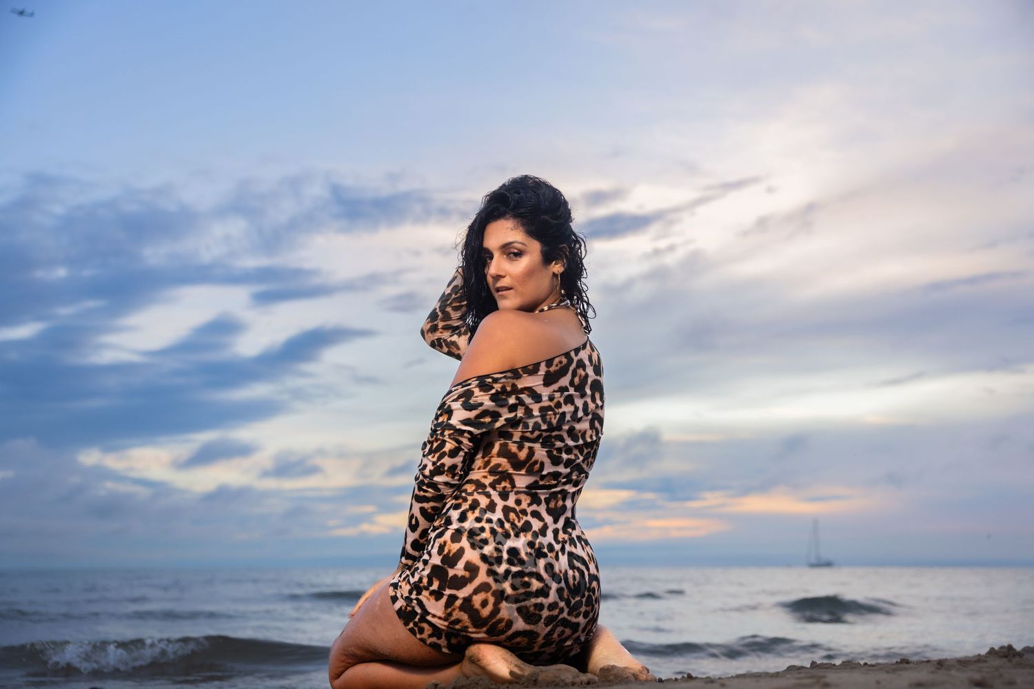 Brunette woman in leopard dress sitting on beach at dusk by ocean.