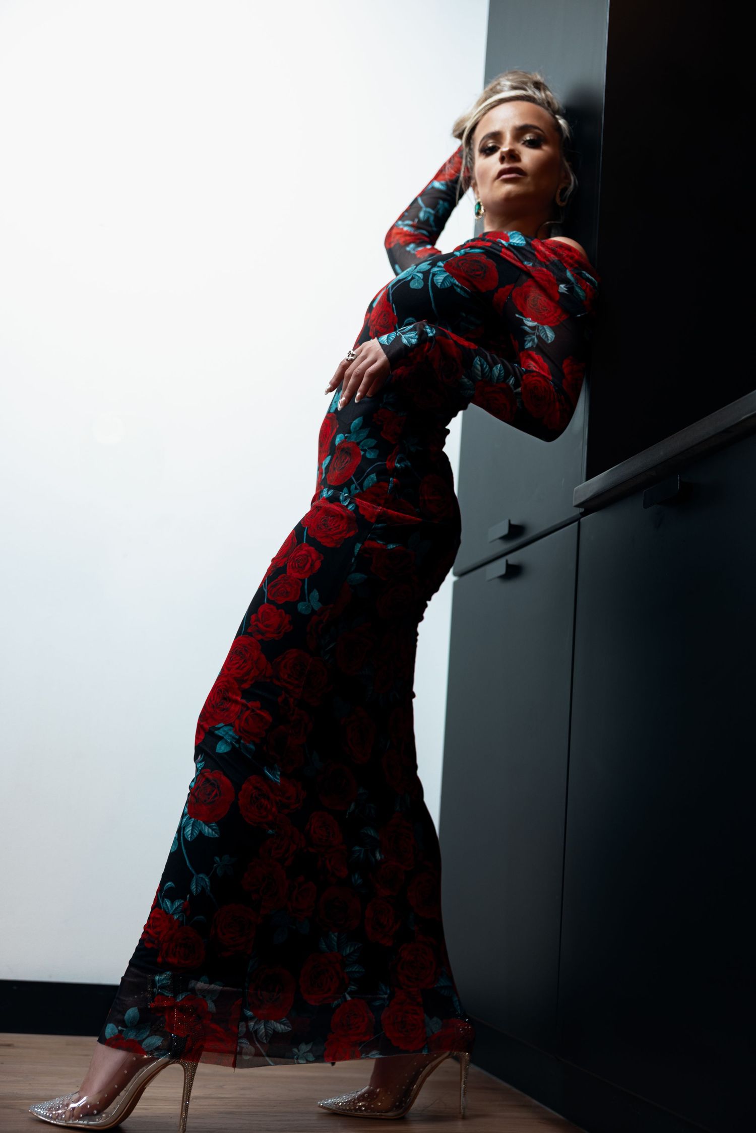 Elegant woman in red rose print maxi dress posing against dark wall in clear heels.