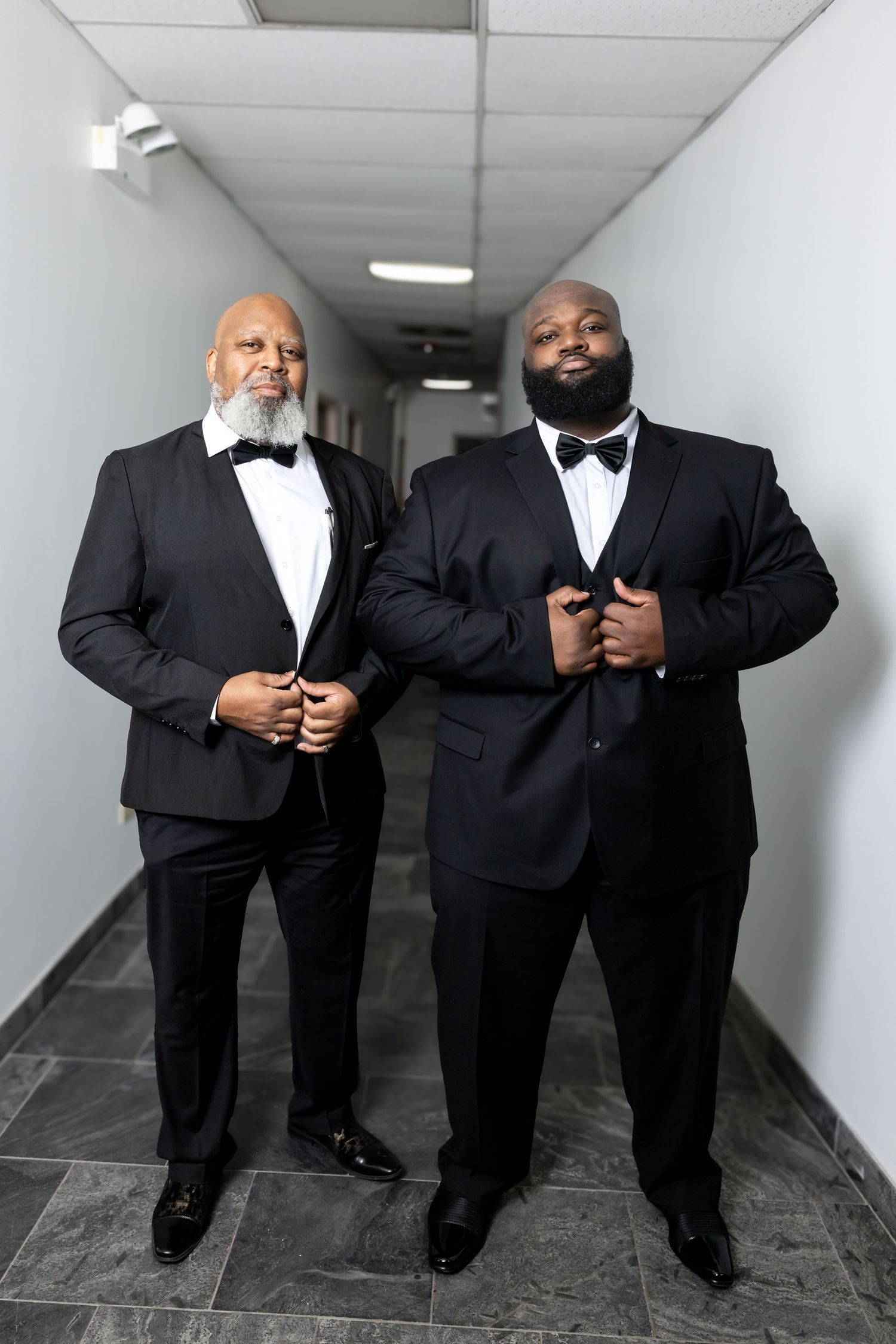 Two men in black tuxedos adjusting their jackets while posing in a well-lit corridor.