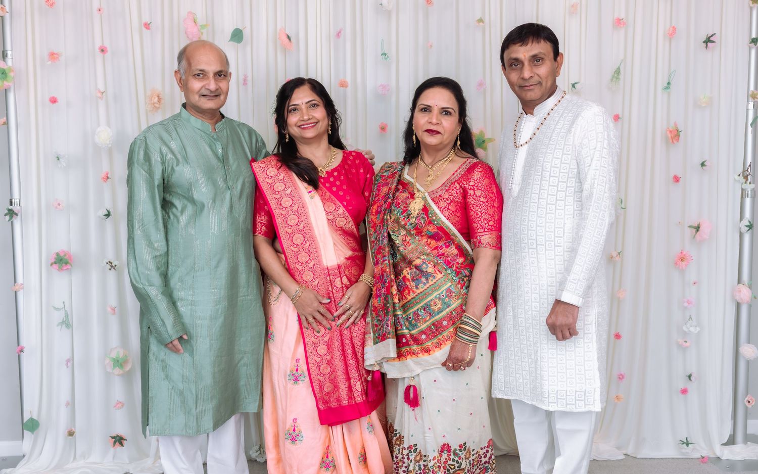 Four Indian adults in traditional festive attire posing together against a white floral backdrop.