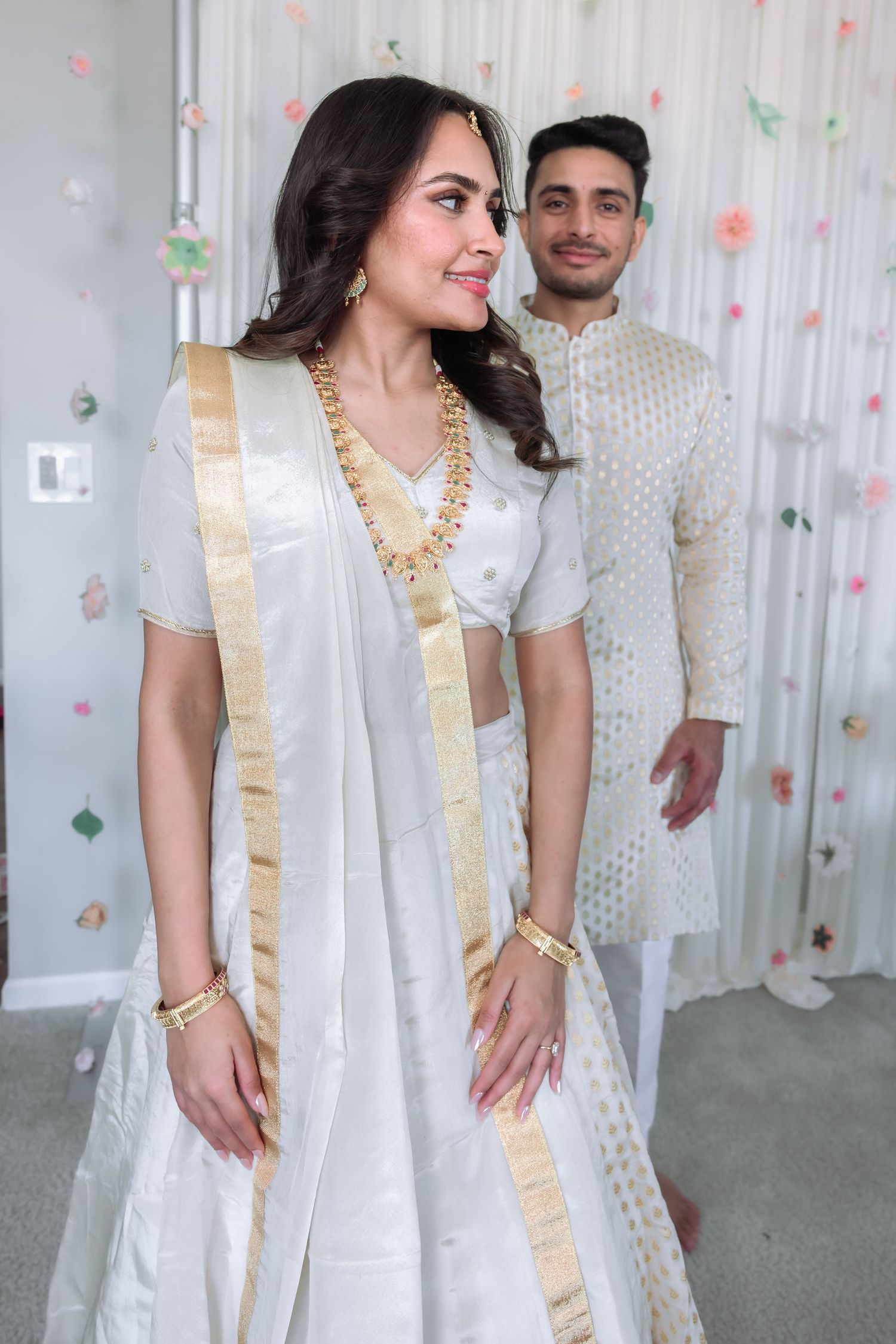 South Asian couple in white traditional Indian wedding attire posing before a floral decorated backdrop.