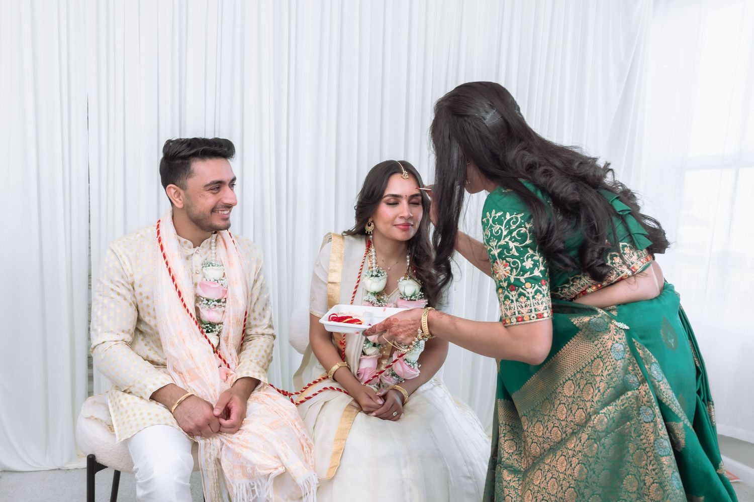 Woman in green saree offering ceremonial plate to smiling Indian bride and groom during wedding ritual.