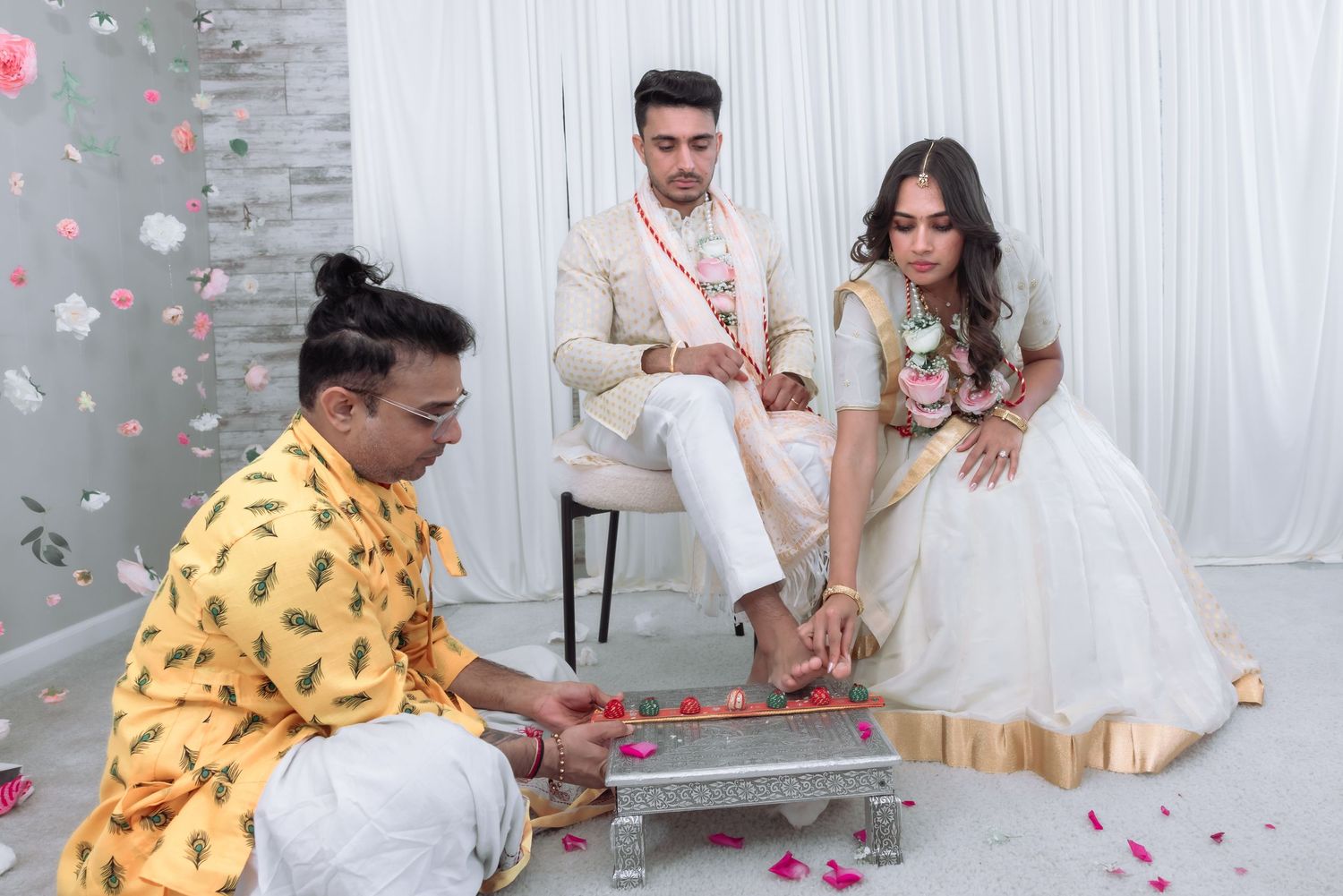 Groom sits on chair as priest performs traditional Hindu wedding foot ritual with bride in white lehenga.