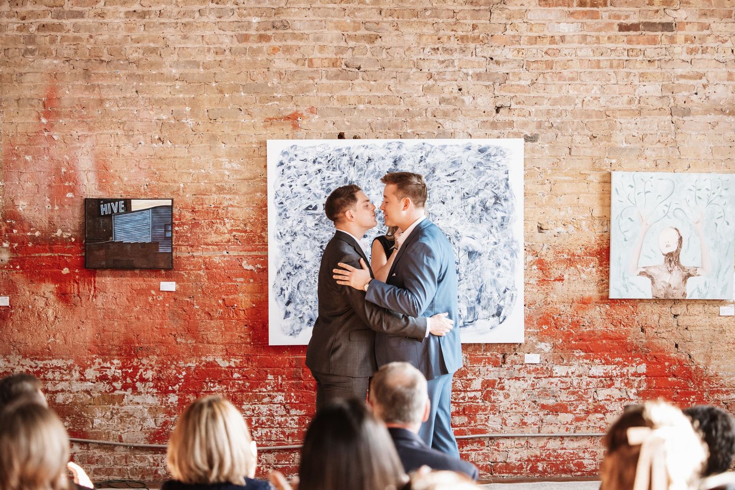 Two grooms share an intimate moment during their wedding ceremony in an art gallery with exposed brick walls.