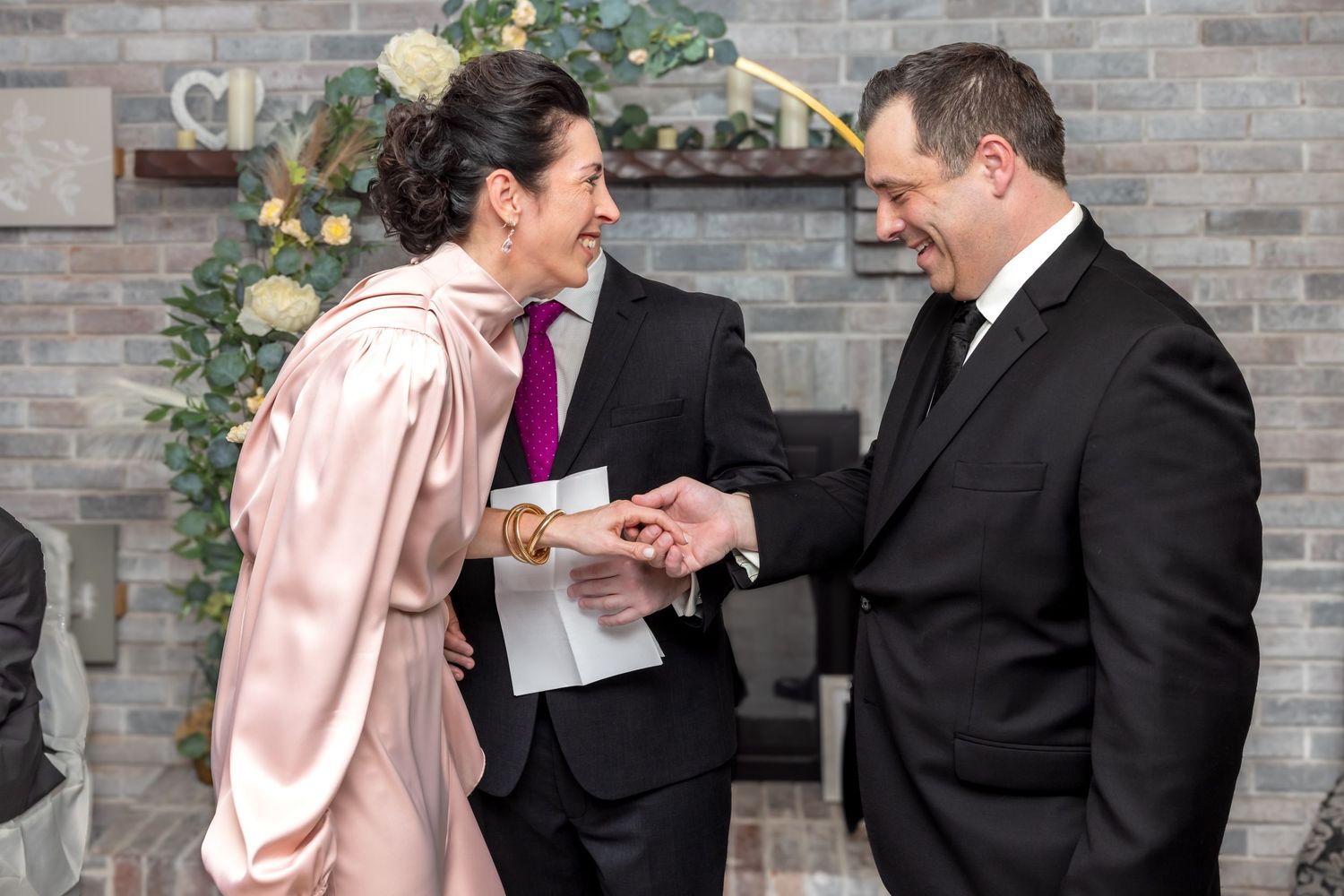 Bride in pink satin gown exchanges rings with groom in black suit at small wedding ceremony.
