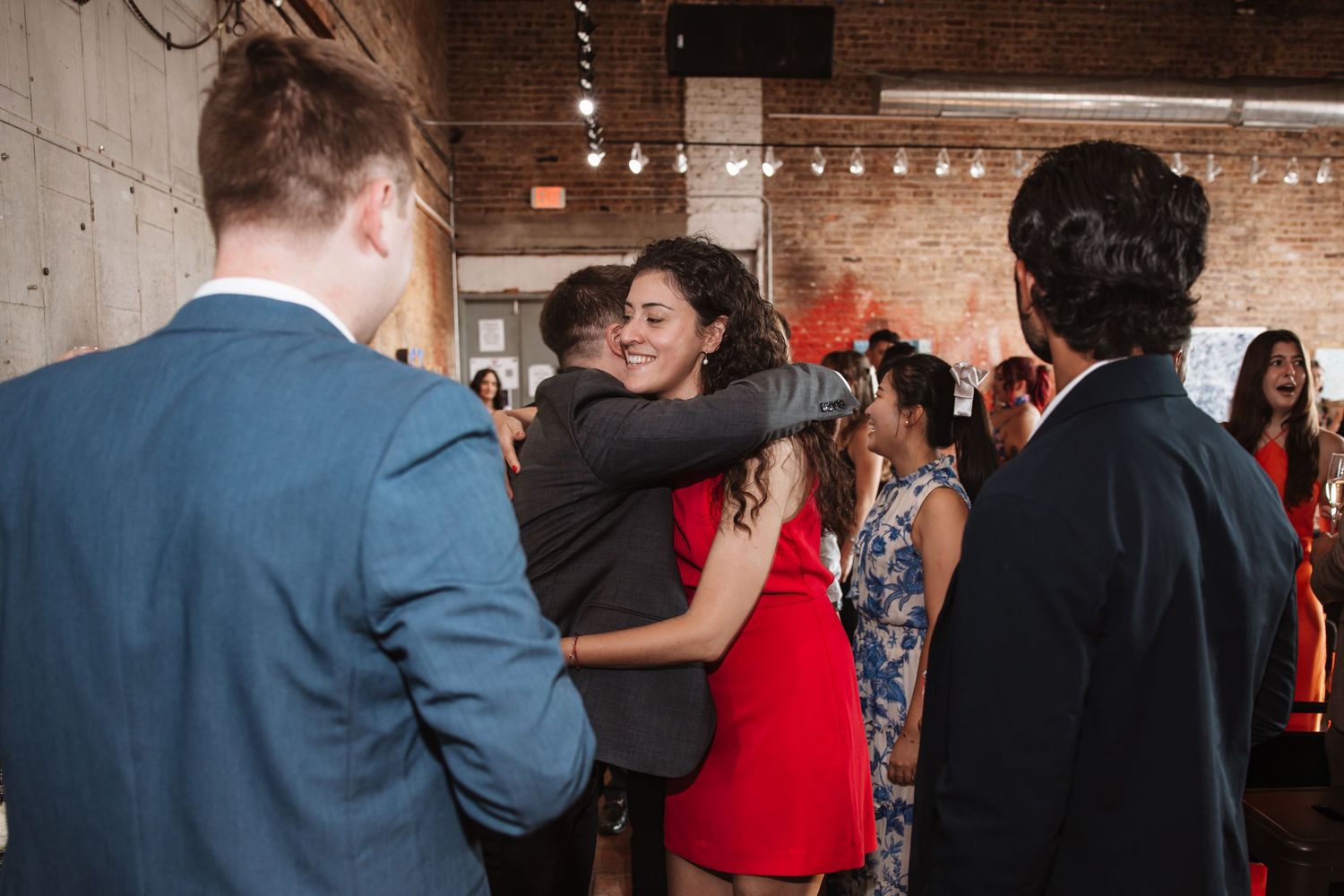 Guests embrace warmly at an indoor social gathering in an industrial-style venue.