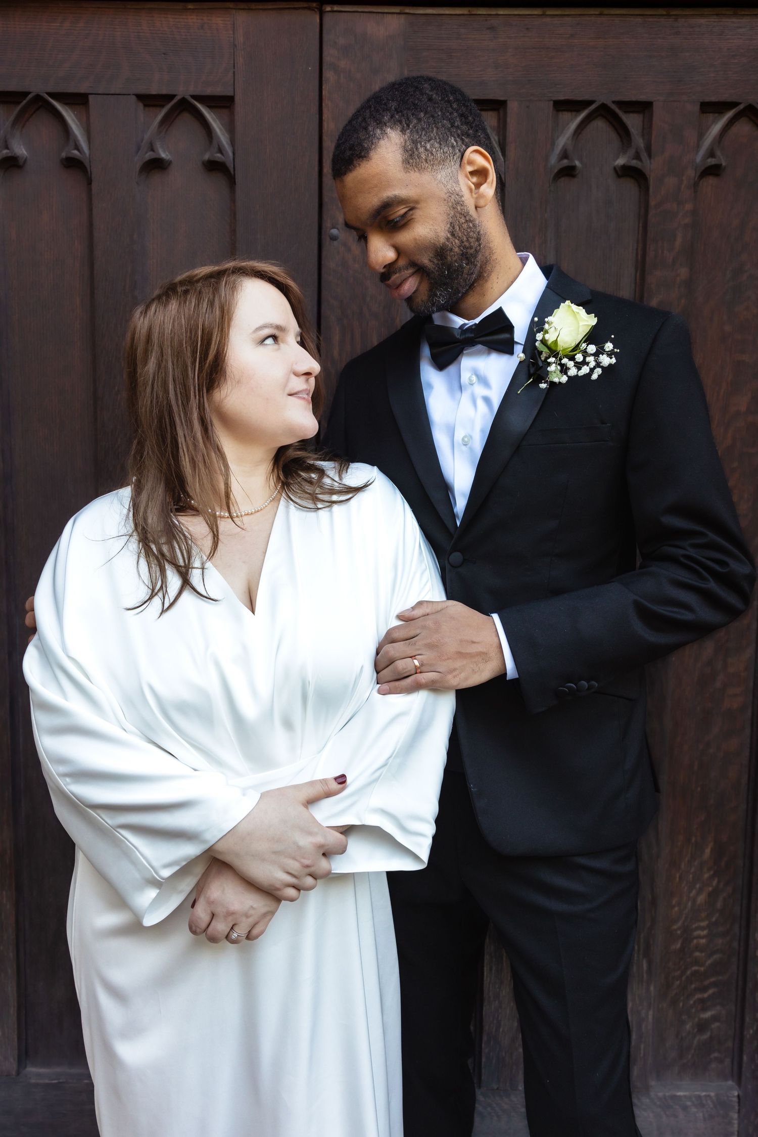 Interracial bride and groom share a loving gaze at church doors.