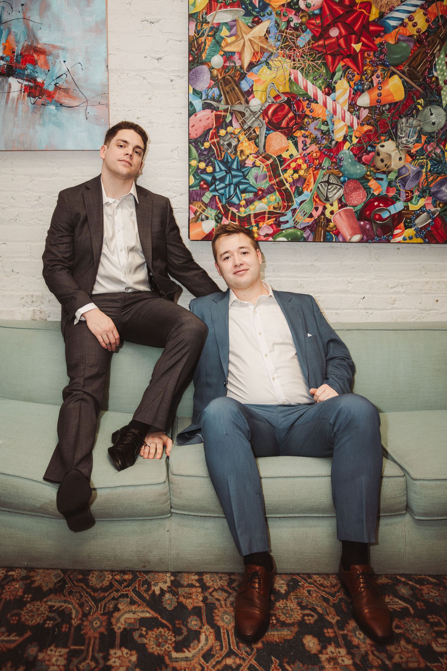 Two stylish young men in suits posed on a mint green sofa with vibrant abstract art backdrop.