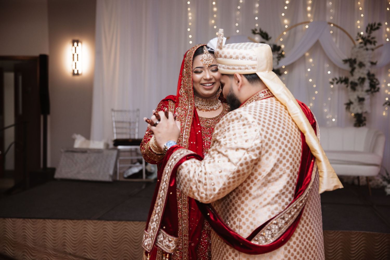 Indian bride and groom share a tender moment in traditional red and gold wedding attire.