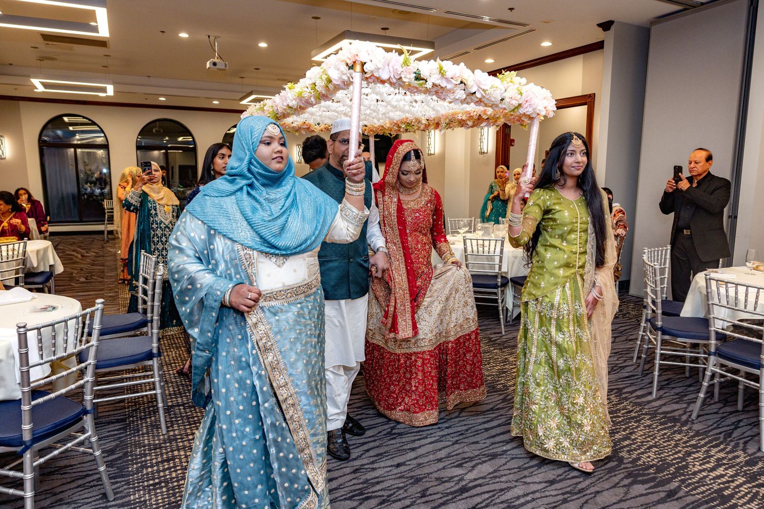 South Asian bride in red bridal attire escorted under floral phoolon ki chaadar at wedding venue.