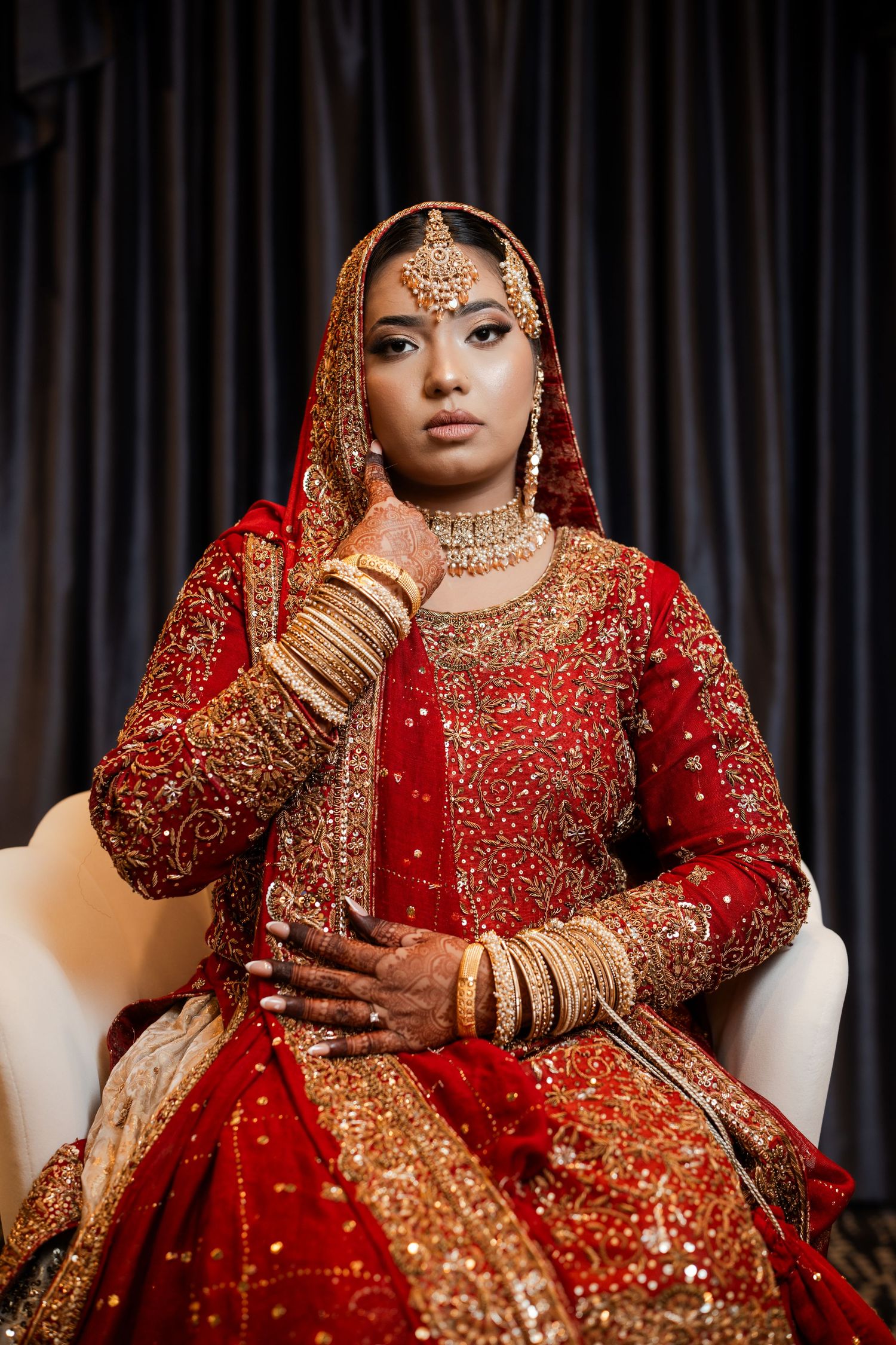 A gorgeous Indian bride sits elegantly in a red and gold embroidered lehenga adorned with gold jewelry.