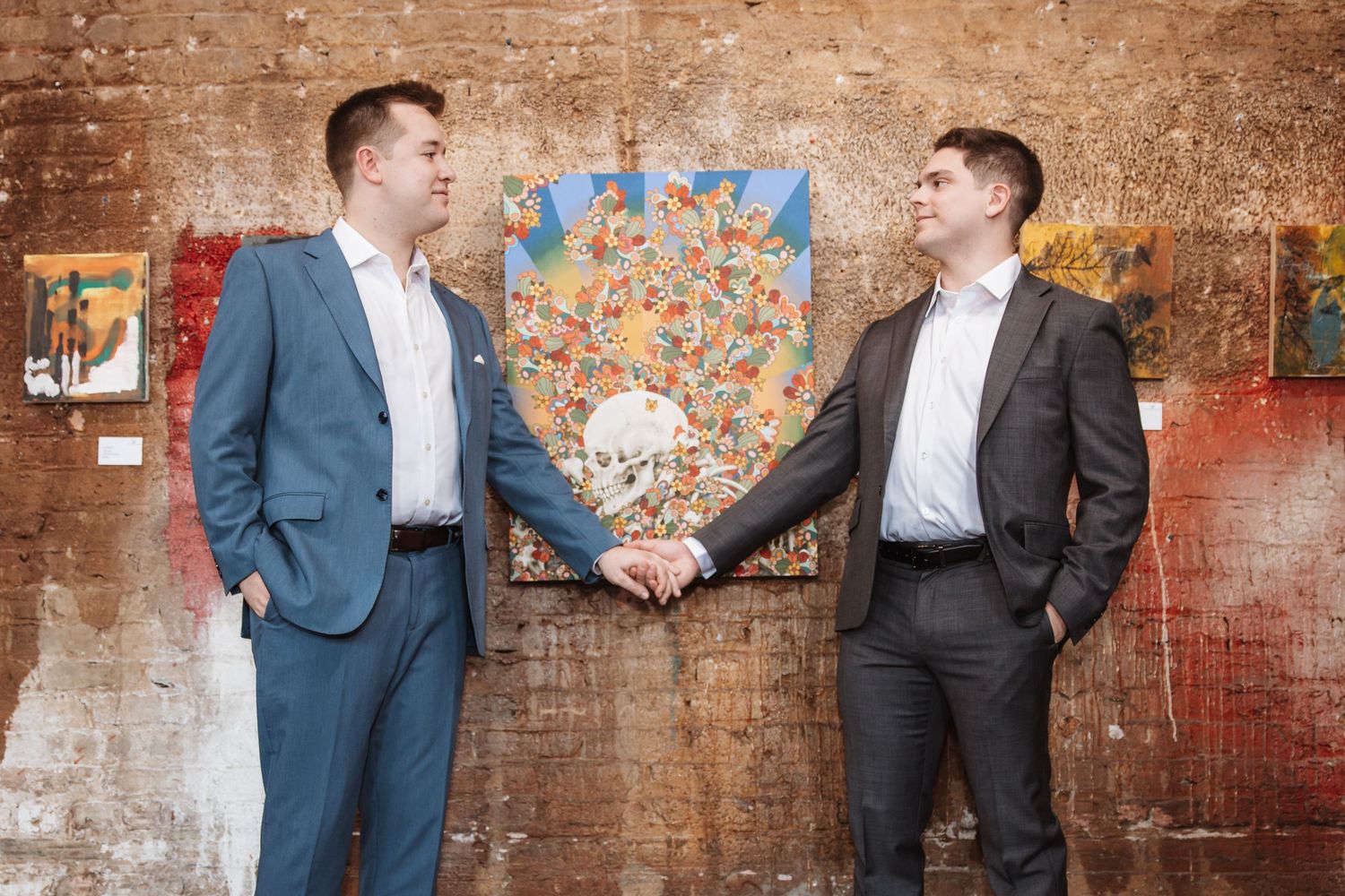 Two smartly dressed men shaking hands while standing before vibrant paintings on a brick wall.