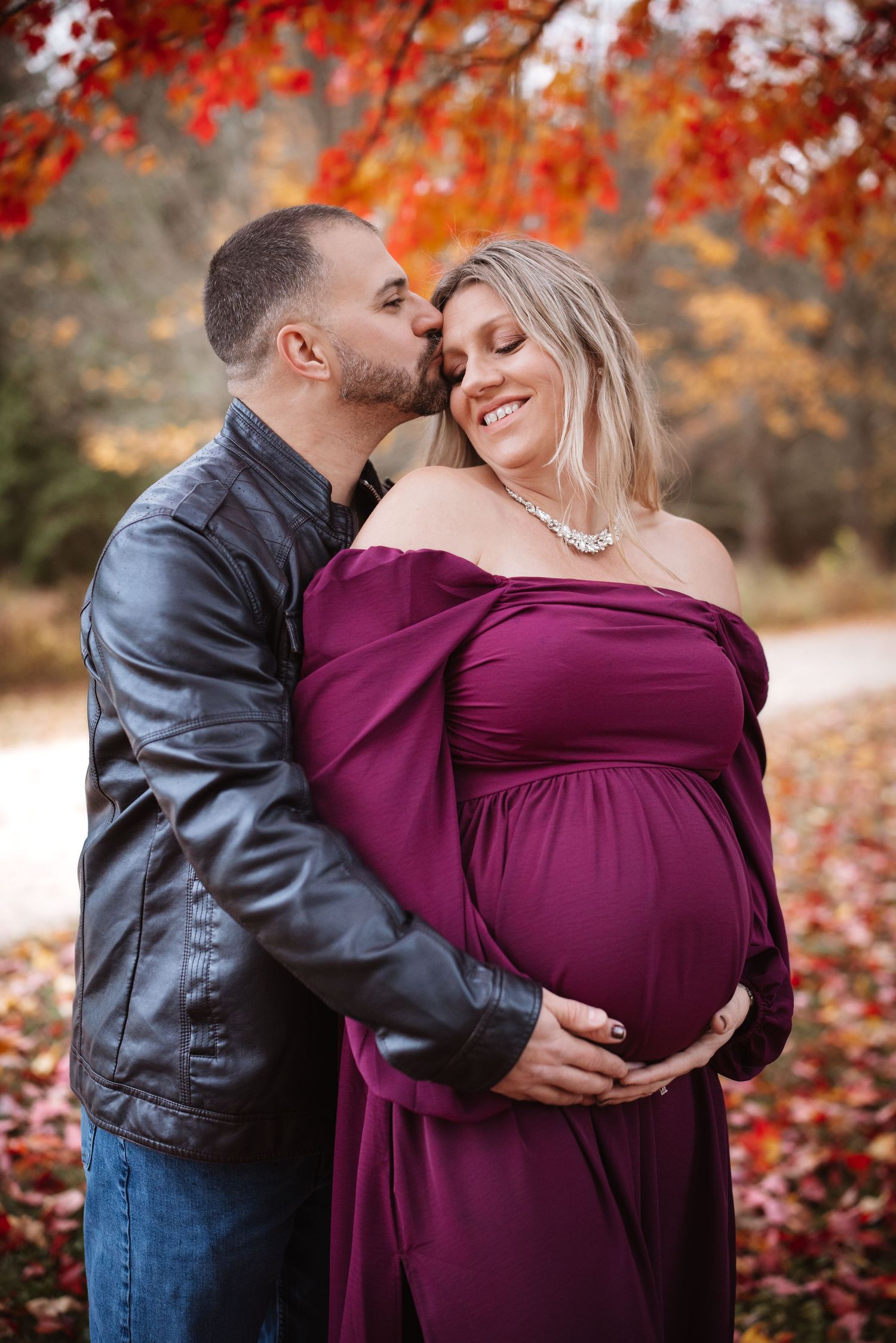 Couple sharing romantic moment outdoors, man kissing pregnant woman's temple in vibrant fall foliage.