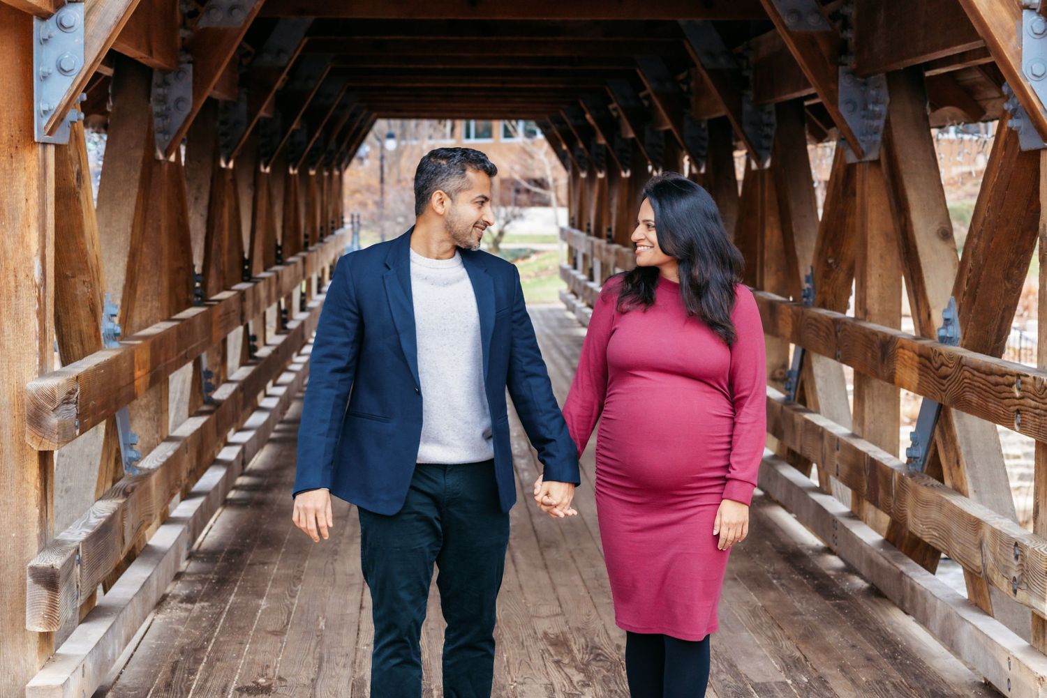 Pregnant couple holding hands walking on a wooden covered bridge, man in navy blazer, woman in burgundy dress.