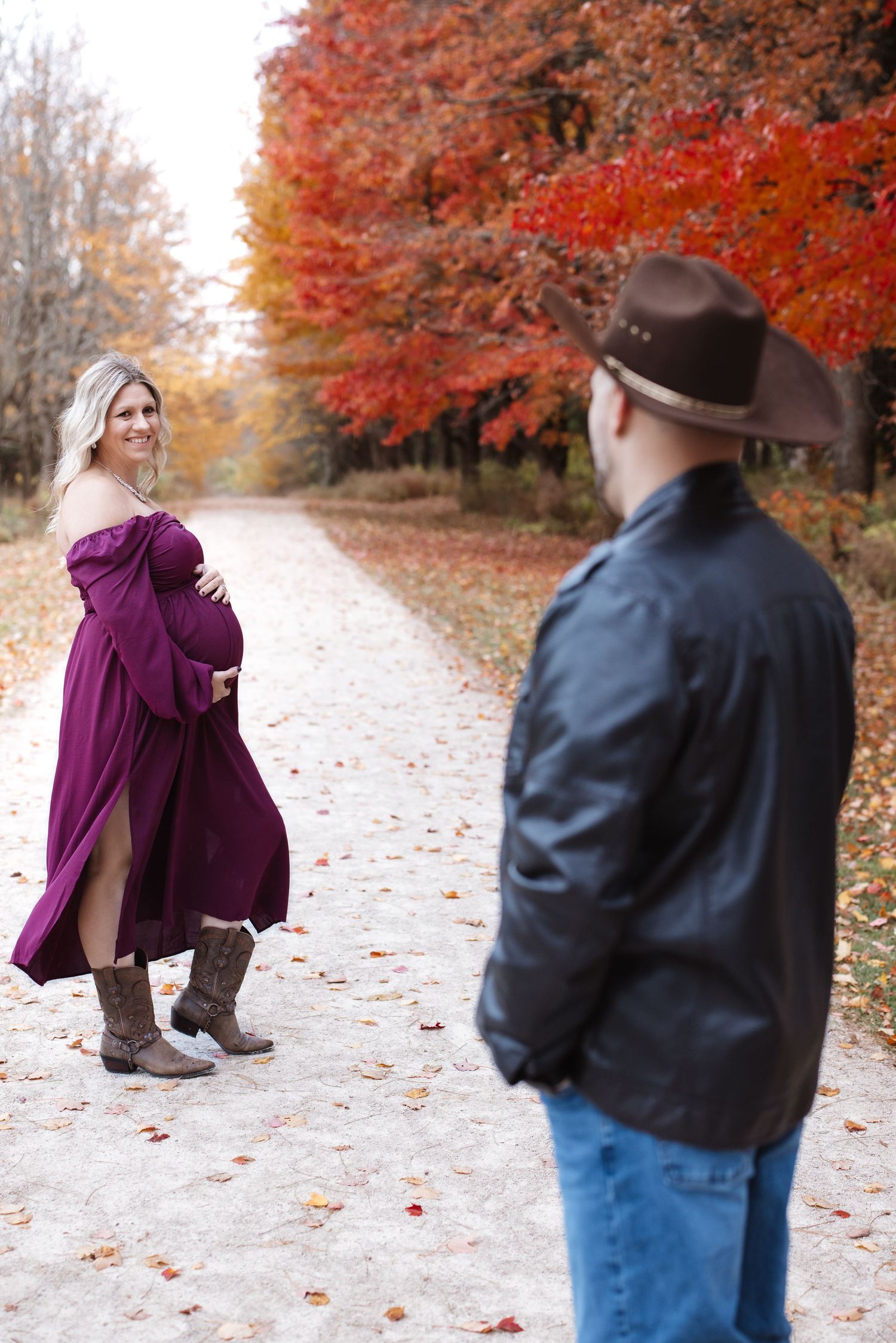 Pregnant woman in burgundy dress smiles at partner on a stunning red and orange autumn path.