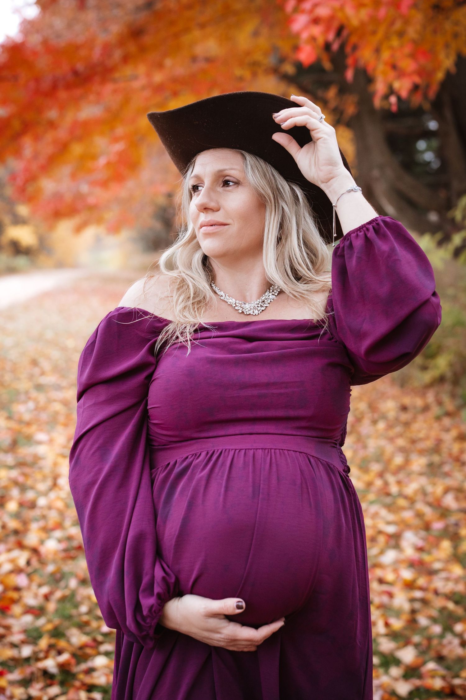 Pregnant woman in purple dress and graduation cap holding baby bump among vibrant autumn foliage.