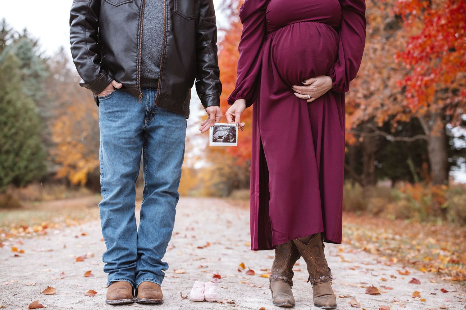Expecting couple holding ultrasound photo with pink baby shoes on autumn park pathway.