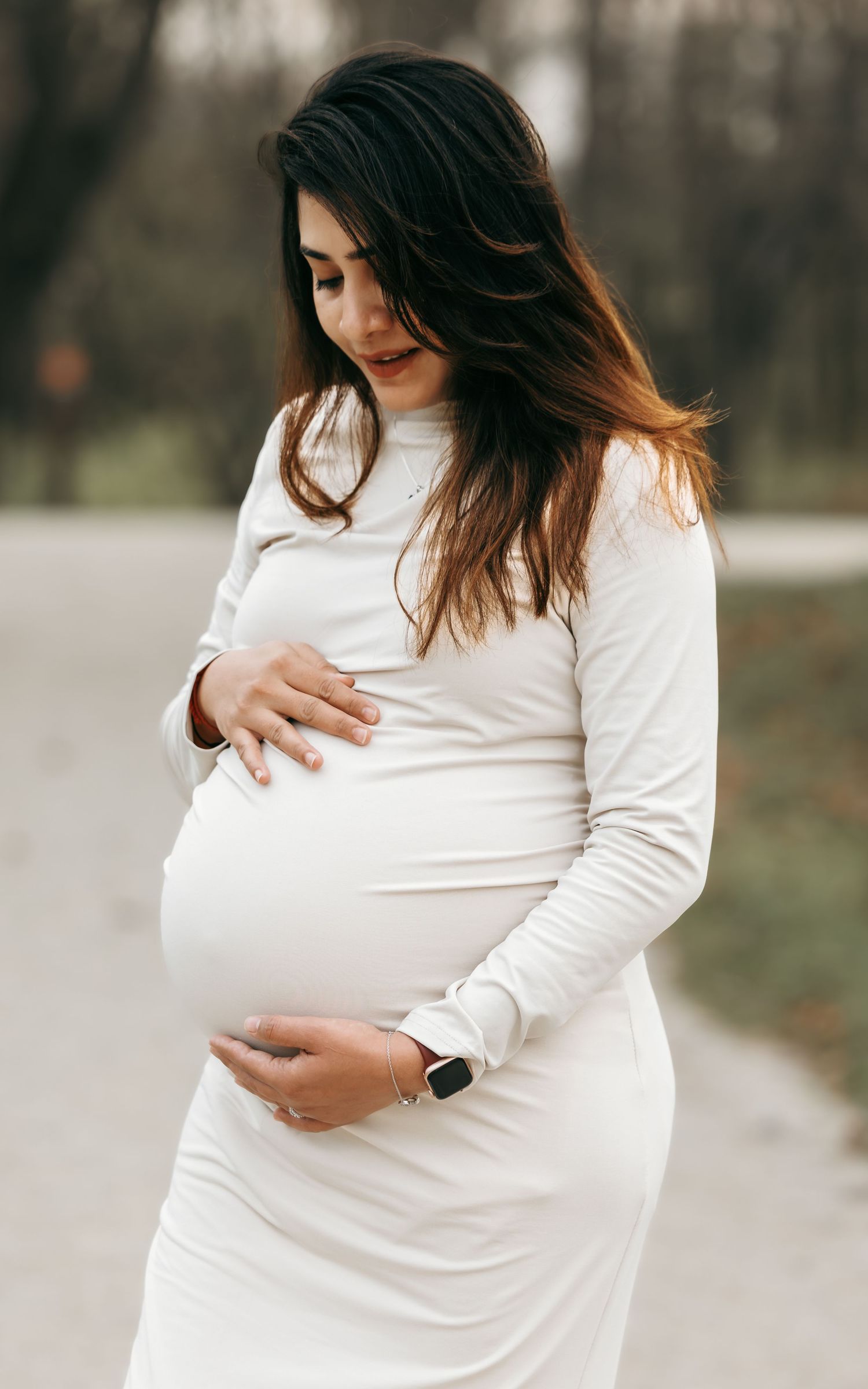 Pregnant woman in white long-sleeve dress cradling her baby bump outdoors in a park setting.