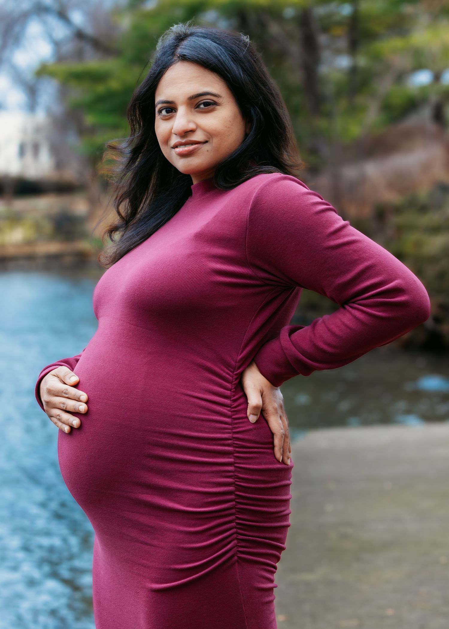 Glowing pregnant woman in burgundy ruched maternity dress smiling, hand on hip near park pond.