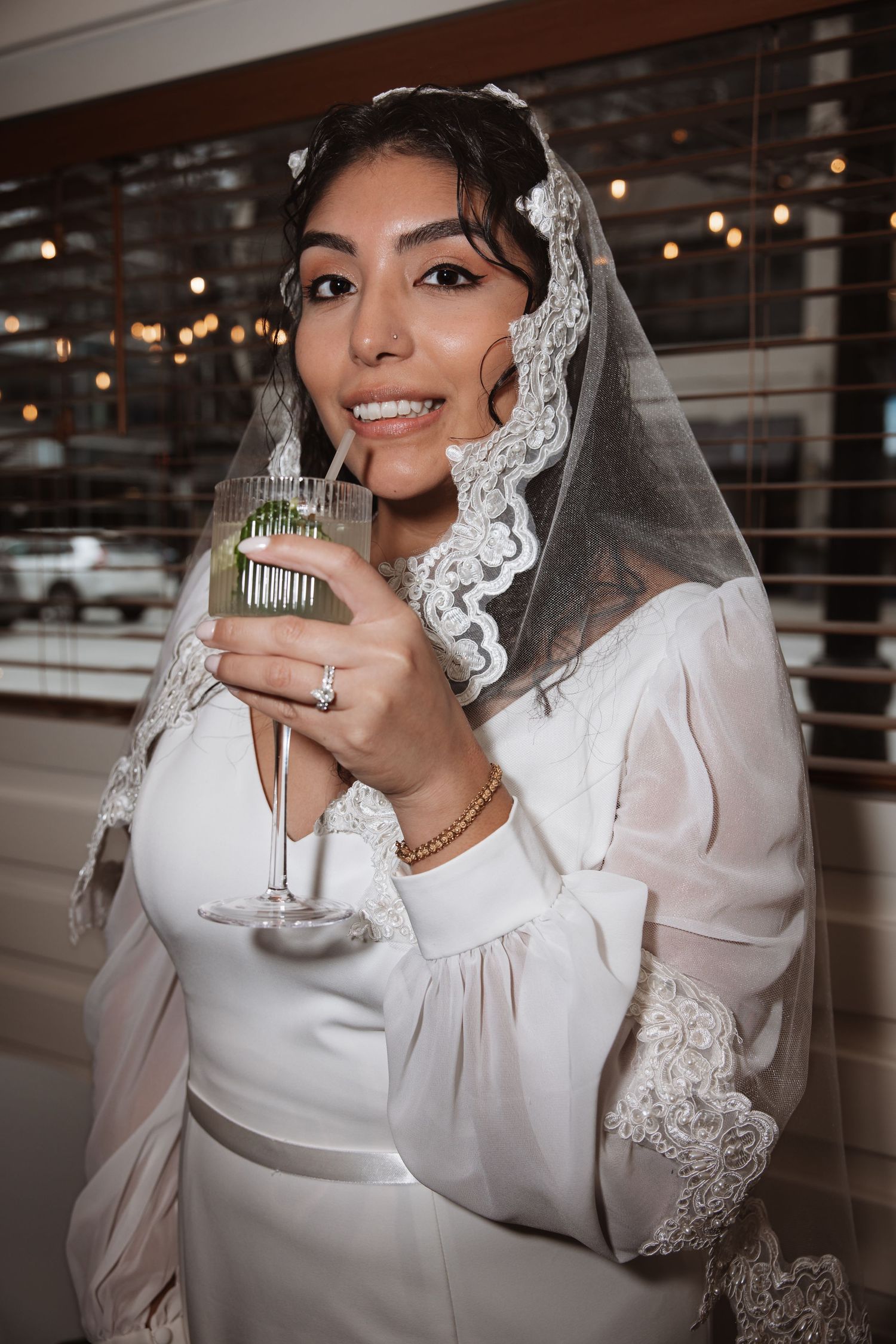 Smiling bride in white wedding dress and lace veil holding a garnished cocktail glass at her wedding reception.