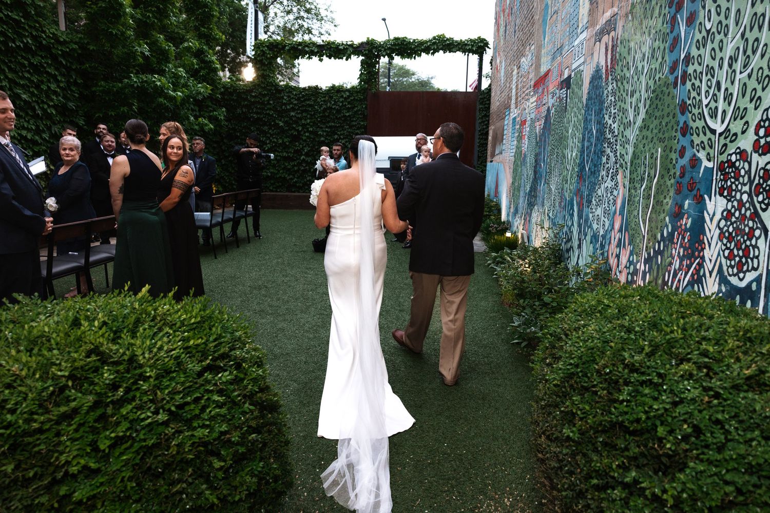A bride in a backless white gown walks toward the altar in a lush outdoor garden wedding ceremony.