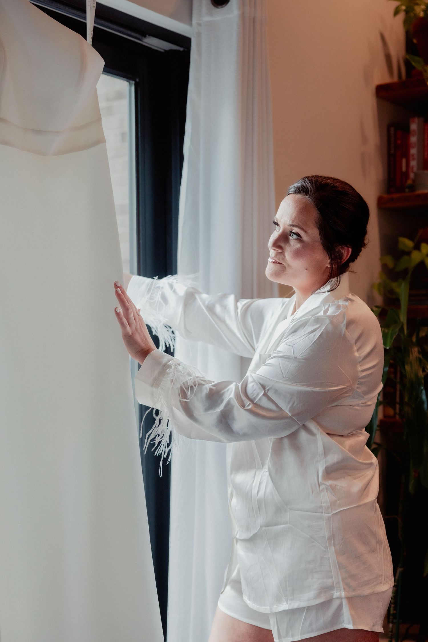 Bride in white feather robe reaching out to touch her wedding gown hanging by window.