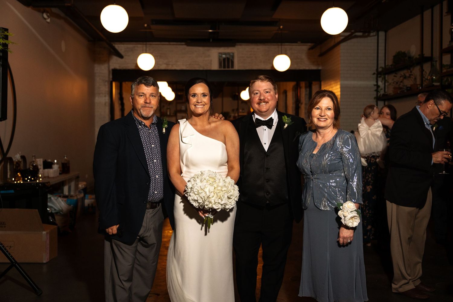 Bride in white gown holds bouquet posing with groom in tuxedo and two family members at wedding reception.